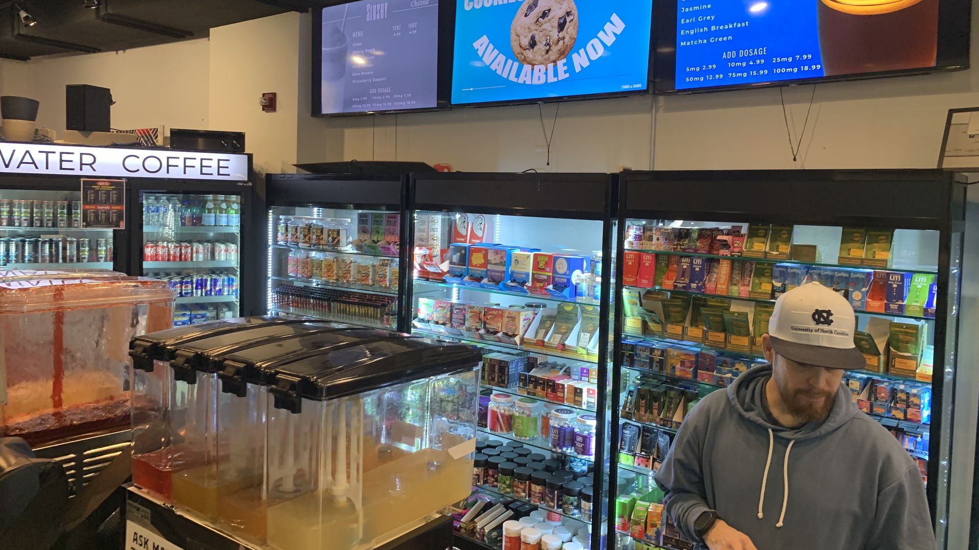 Man wearing a University of North Carolina cap and blue hoodie stands in front of refrigerated shelves stocked with various products in a store with digital menu boards above and drink dispensers in front.