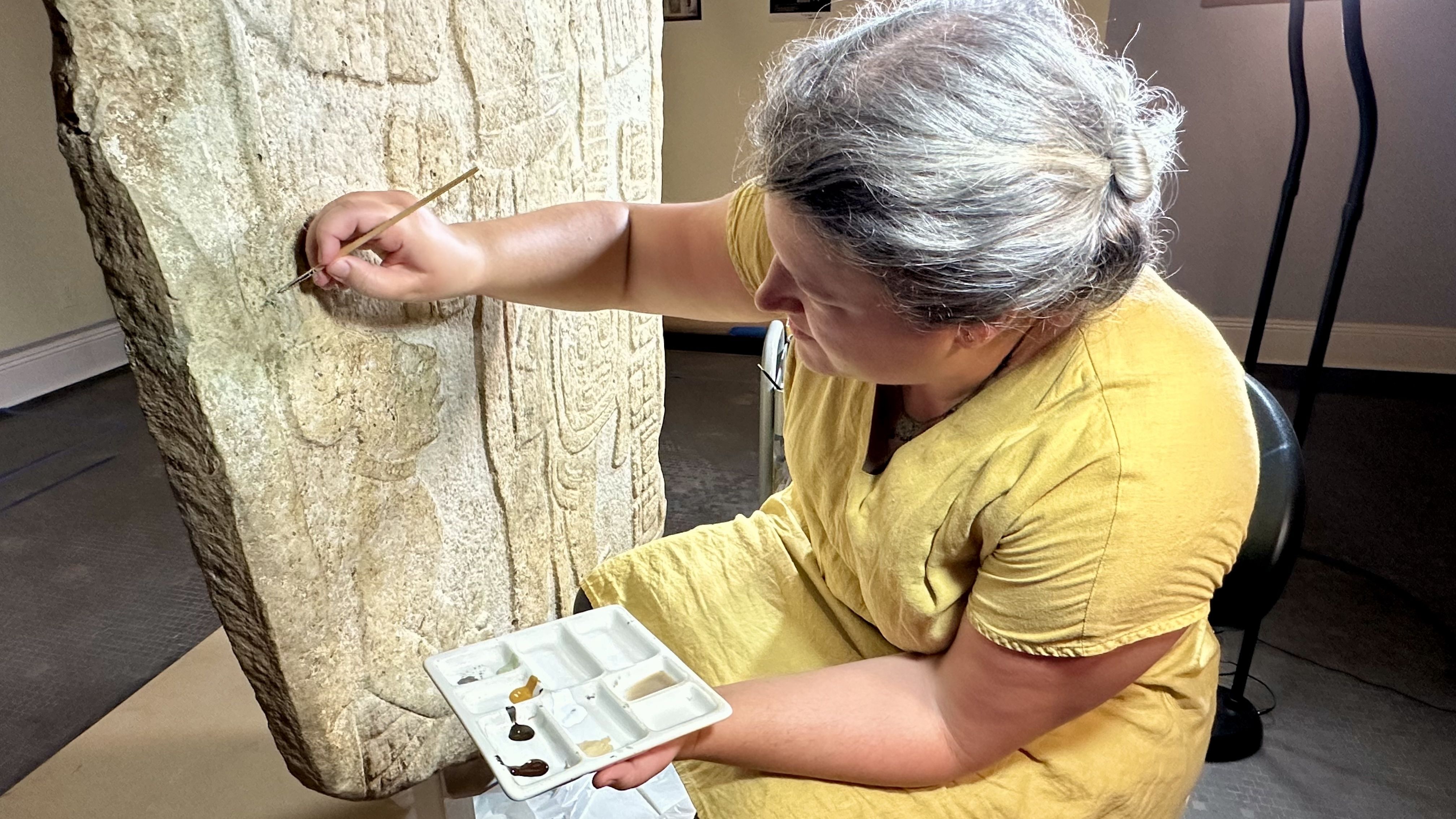 A woman in a yellow tunic sits beside a carved stone stele, carefully painting its engravings with a thin brush; a white palette with browns rests on their lap.