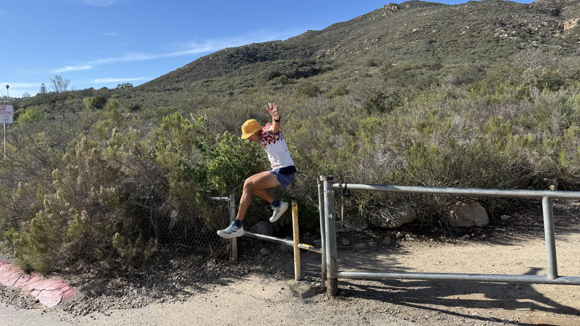 Person wearing a yellow helmet and white shirt with red pattern balances on a metal railing in a dry, scrubby hillside; rocky ridge under a clear blue sky.