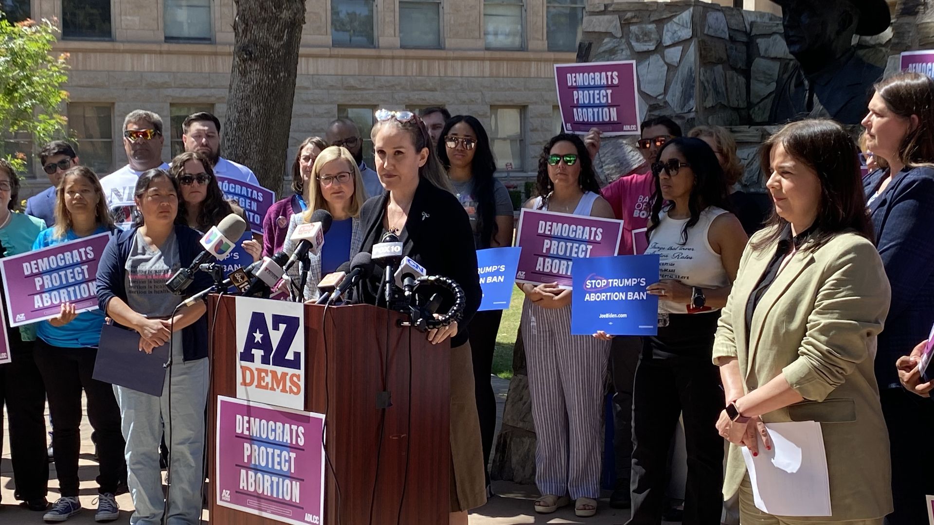 A woman stands at a lectern covered in microphones surrounded by people holding signs.