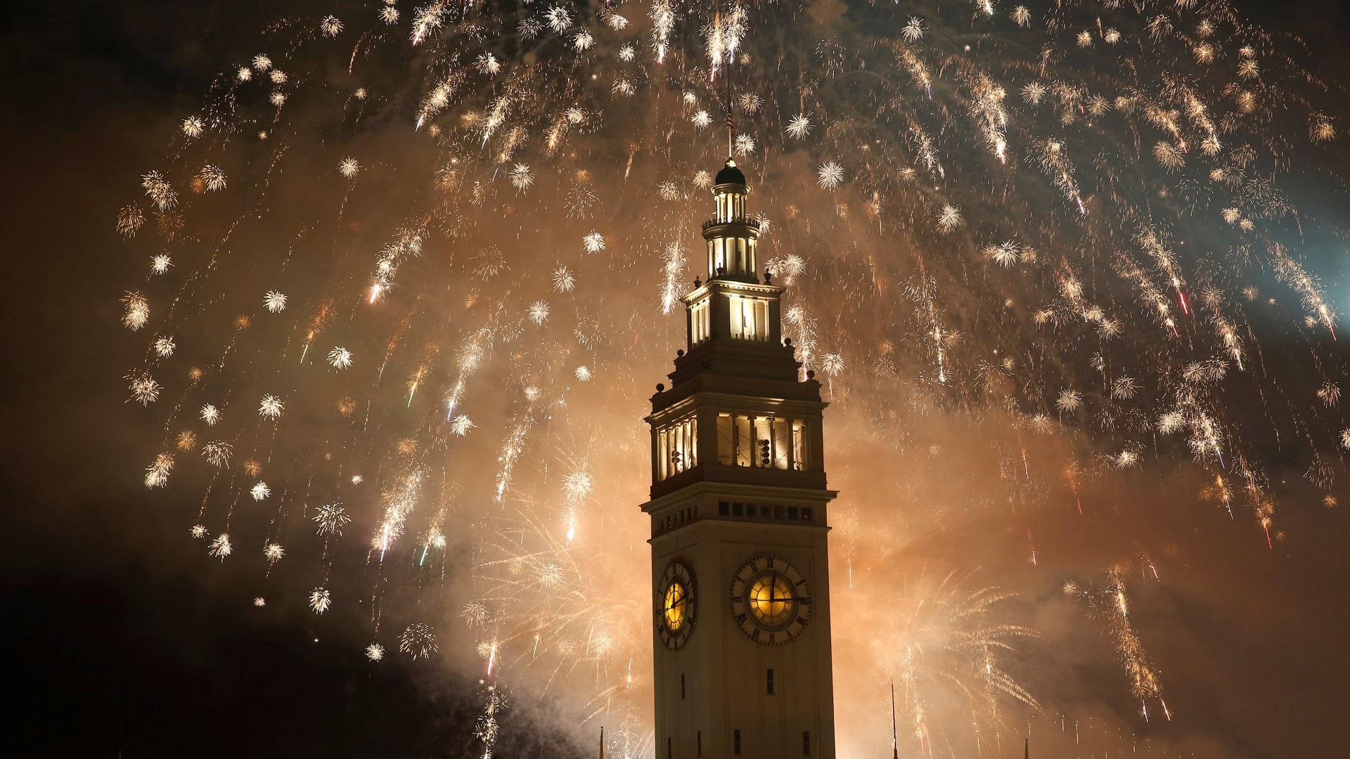 Clock tower illuminated at night with golden fireworks bursting in the sky behind, creating a bright and festive atmosphere.