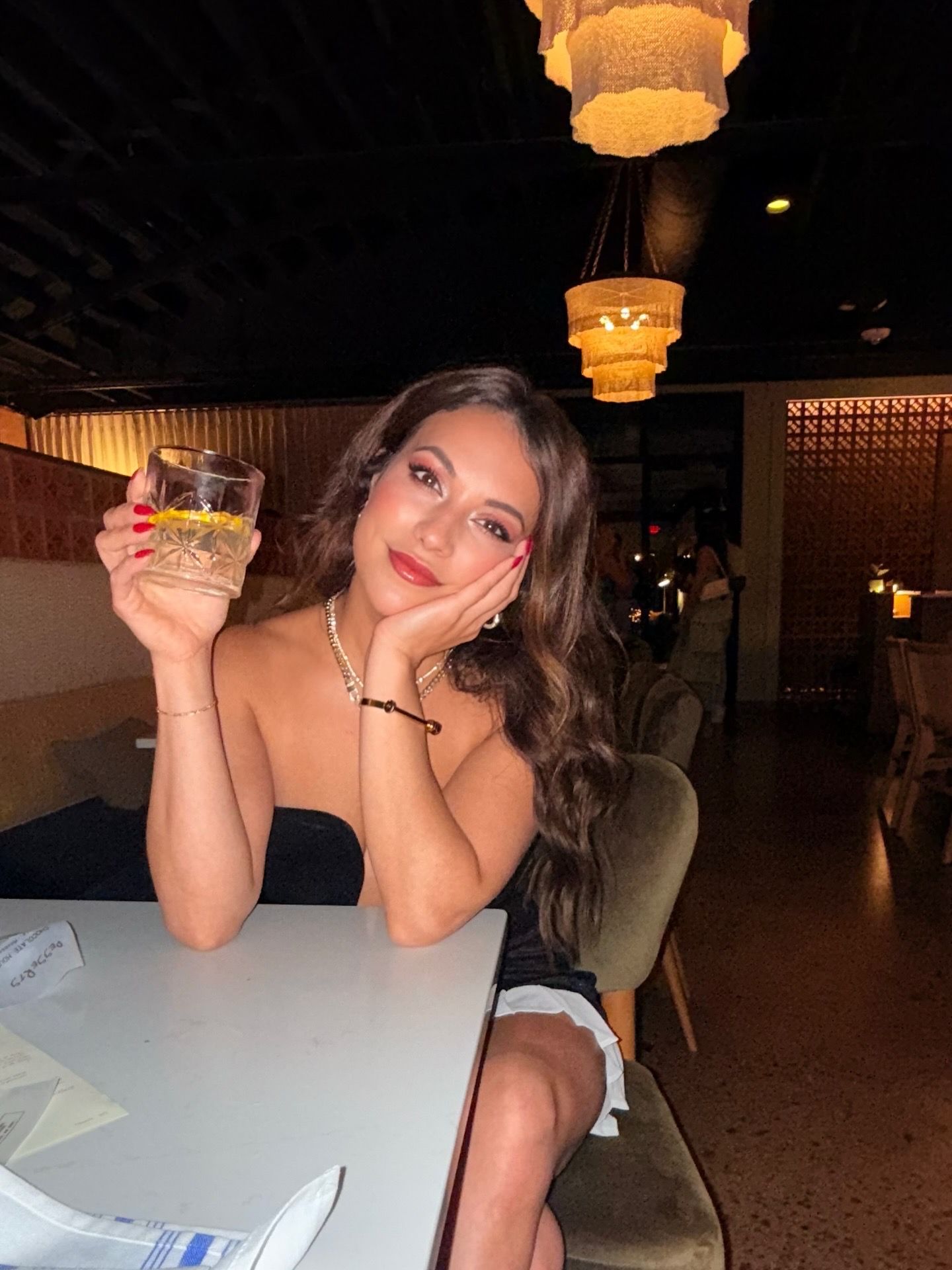 A woman with long wavy brown hair sits at a table in a dimly lit restaurant, wearing a black strapless dress and holding a glass with a lemon slice, smiling with red nails and makeup.