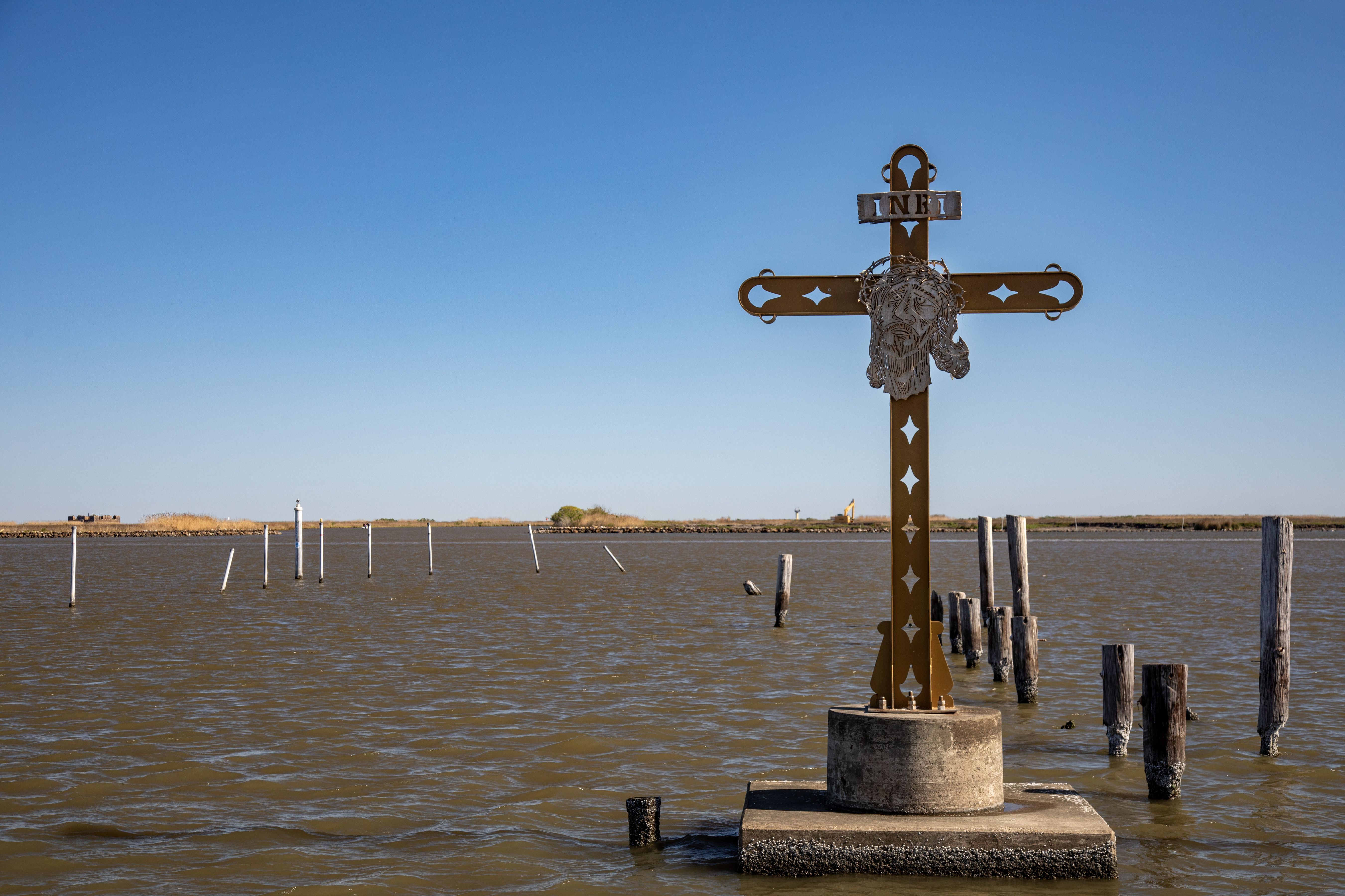 Rust-colored cross with a metal face and crown of thorns, inscribed INRI, stands on a concrete base in murky brown water with wooden posts and clear blue sky in the background.