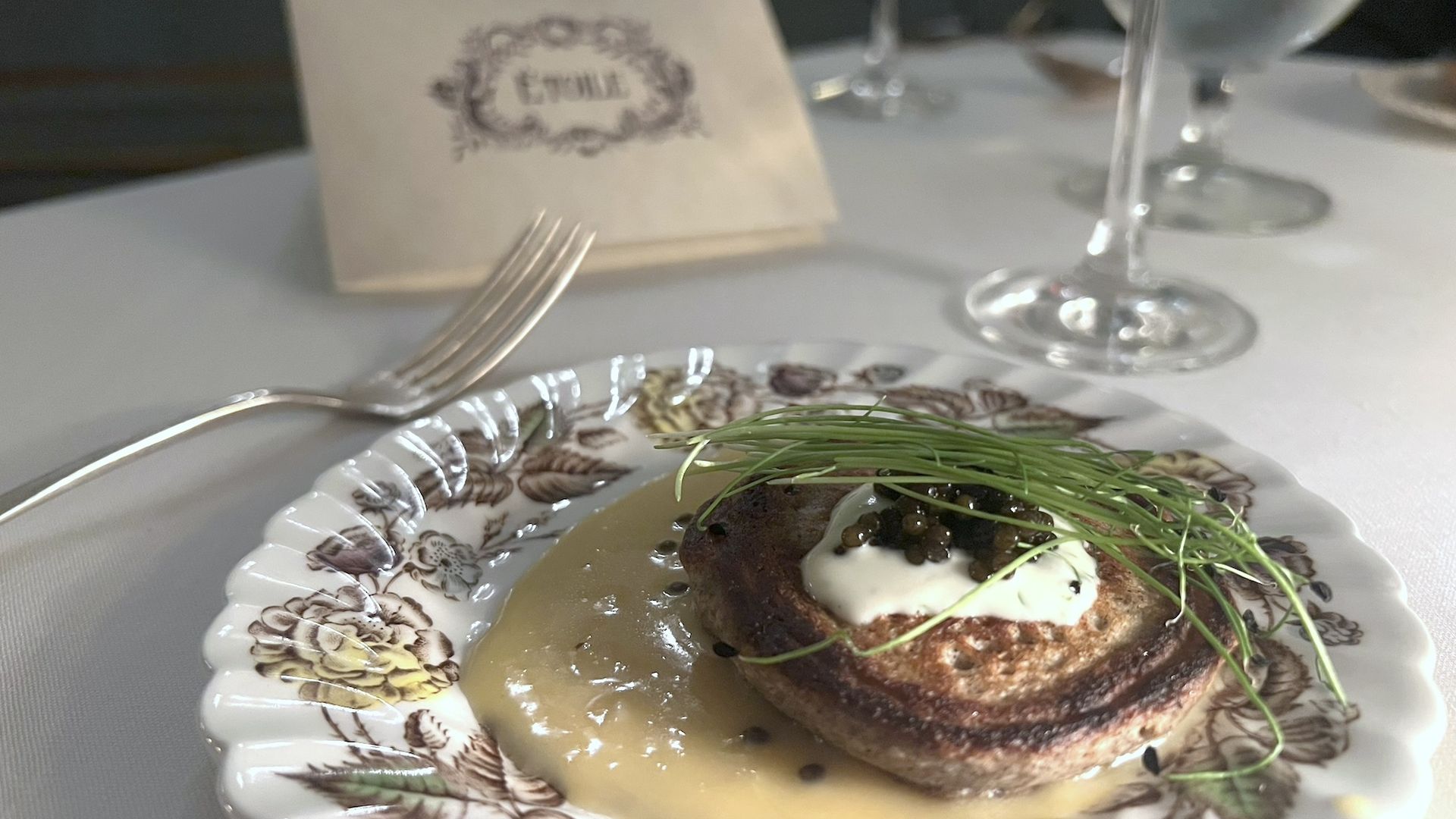 A close-up ohoto of a small pancake-like dish served with caviar on a vintage-looking plate. A paper placard on the table behind it says Etoile.
