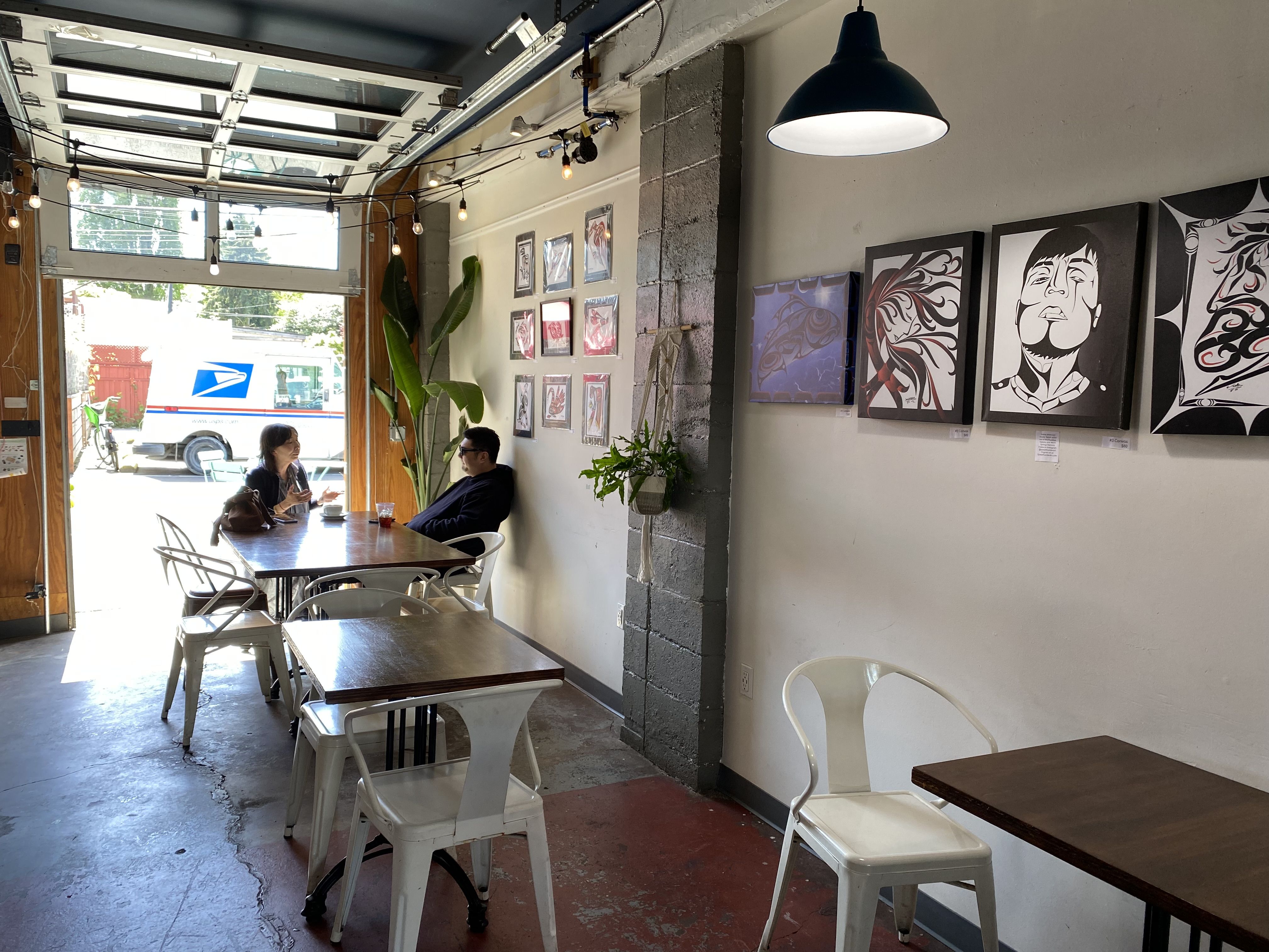 Closer photo of two people talking at coffee shop table with an open garage-style door behind them, art on walls.