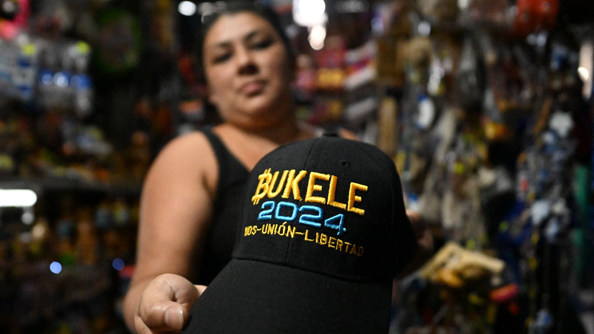 A woman who is blurred in the background holds a hat with the name of Bukele and the numbers 2024