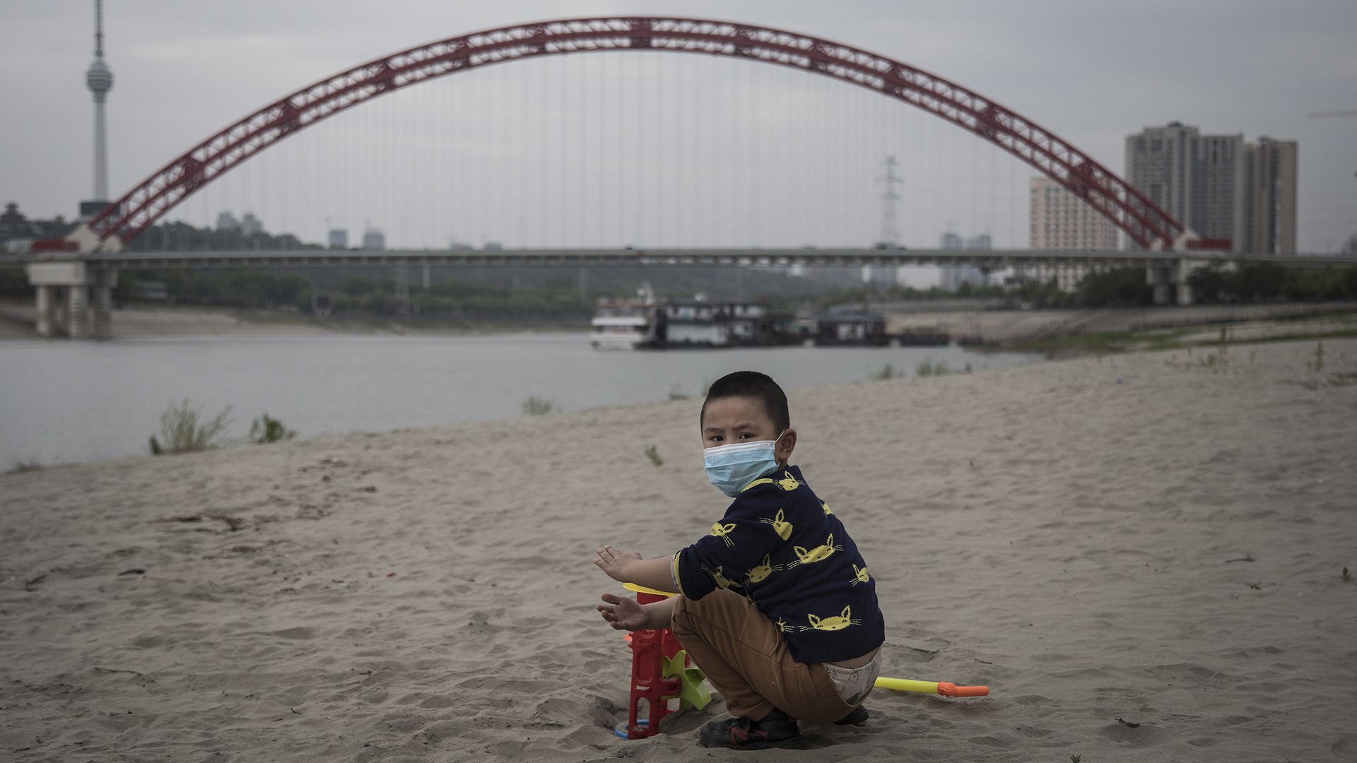 In this image, a boy sits on a beach wearing a face mask