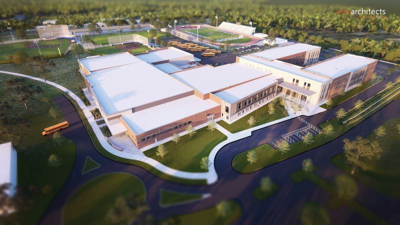 Aerial view of a large modern school campus with white rooftops, multiple rectangular buildings, a football field with bleachers, school buses, and surrounding roads and trees.