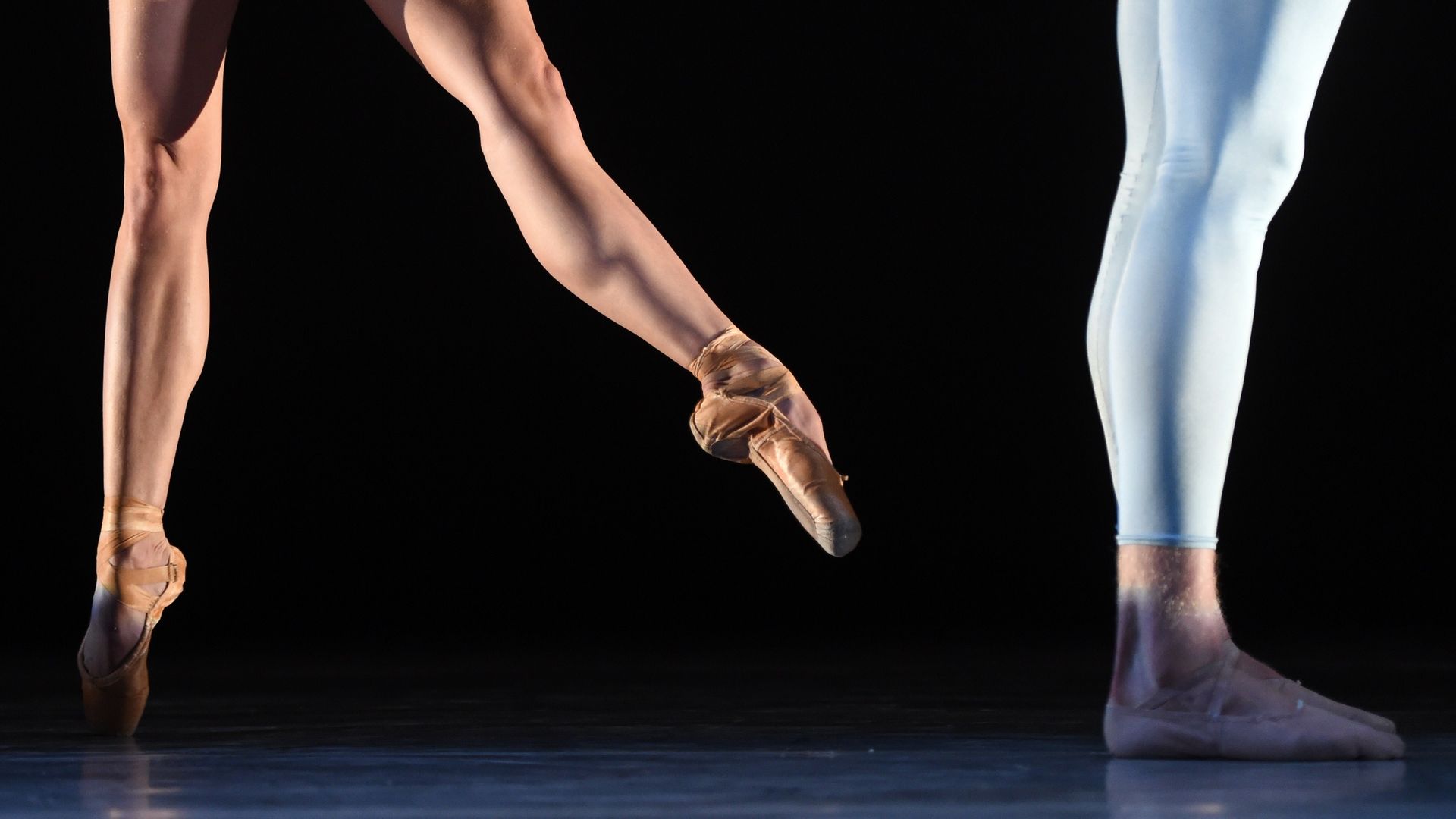 Close-up of two ballet dancers' legs on stage, one on pointe with tan ballet shoes, the other wearing light blue leggings with pink ballet slippers, against a dark background.
