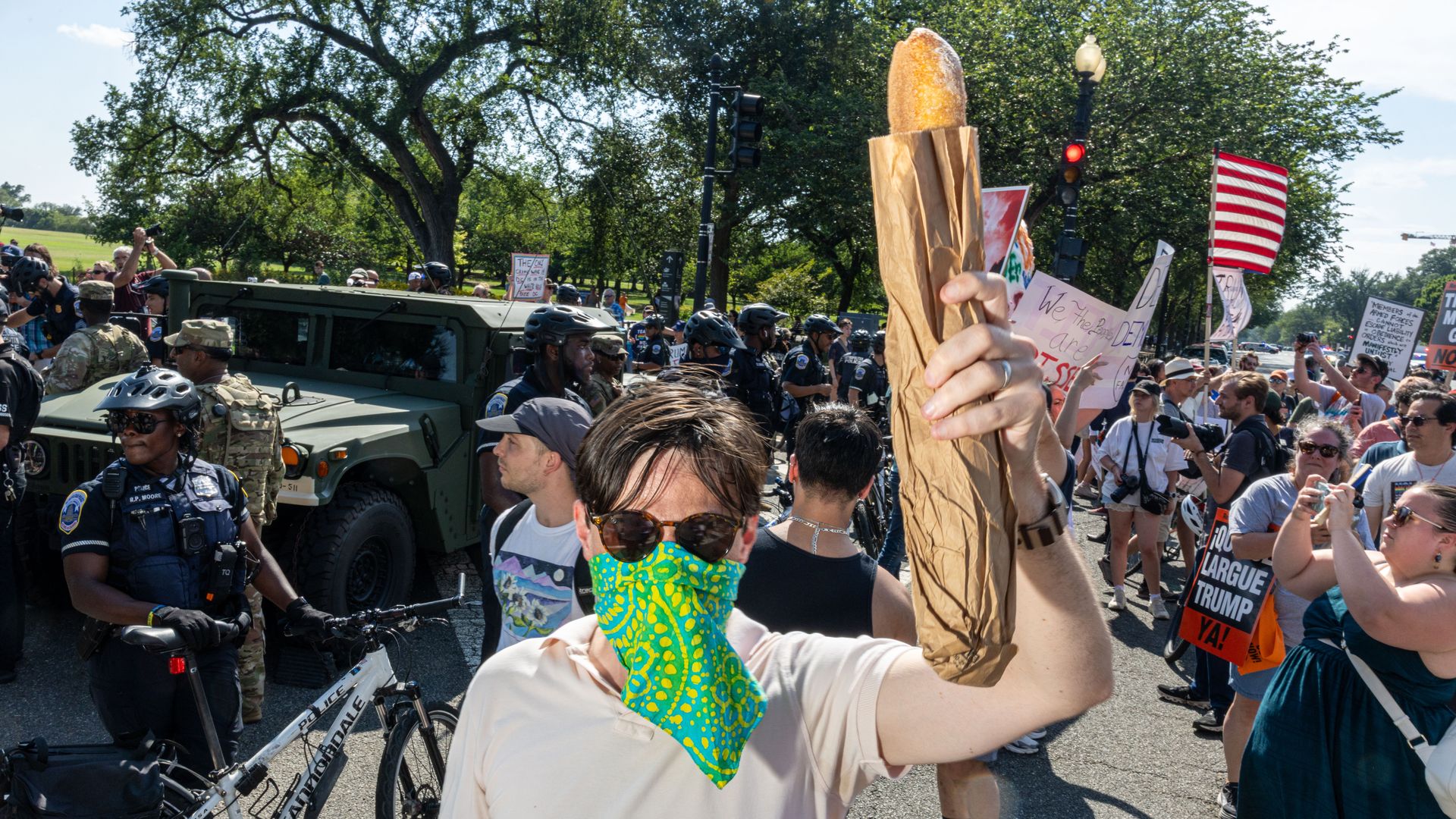 Protester wearing sunglasses and a green patterned face covering holds a wrapped baguette amidst a crowd with signs and police officers with bicycles in a street protest.