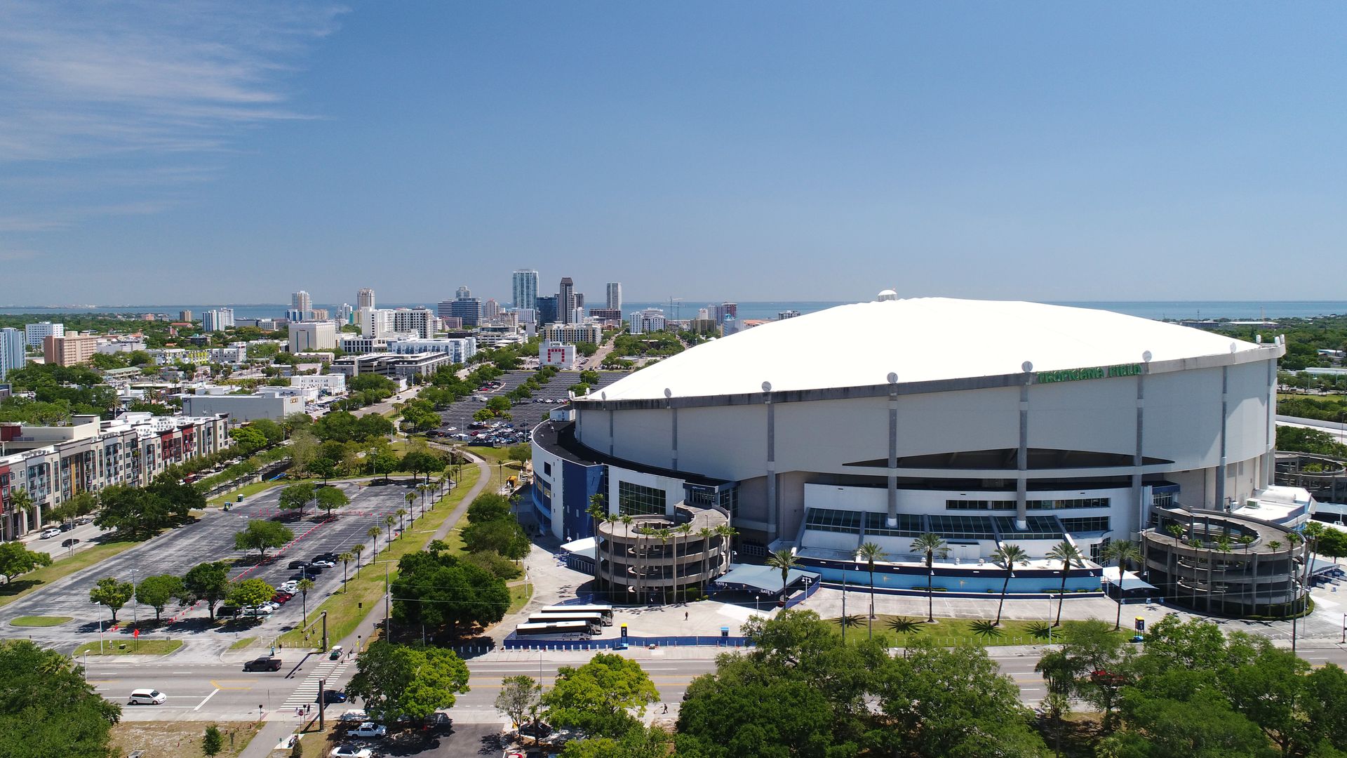 Aerial view of Tropicana Field stadium with its white domed roof on a sunny day, palm trees around it, a parking lot, and the city skyline in the background under a clear blue sky.