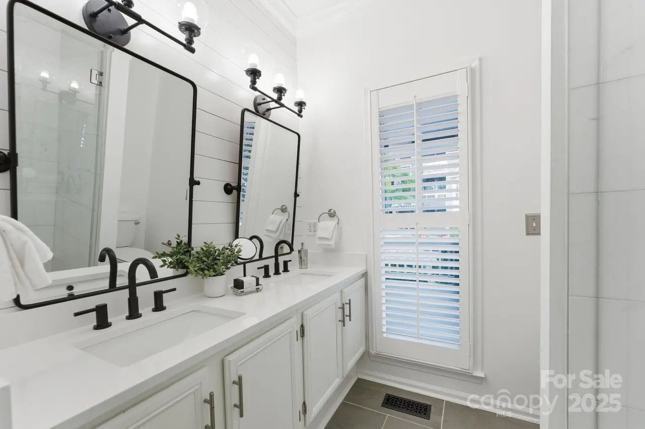 Bright white bathroom with double sinks, black fixtures, two rectangular mirrors, light fixtures above, potted plant, and a window with white plantation shutters.