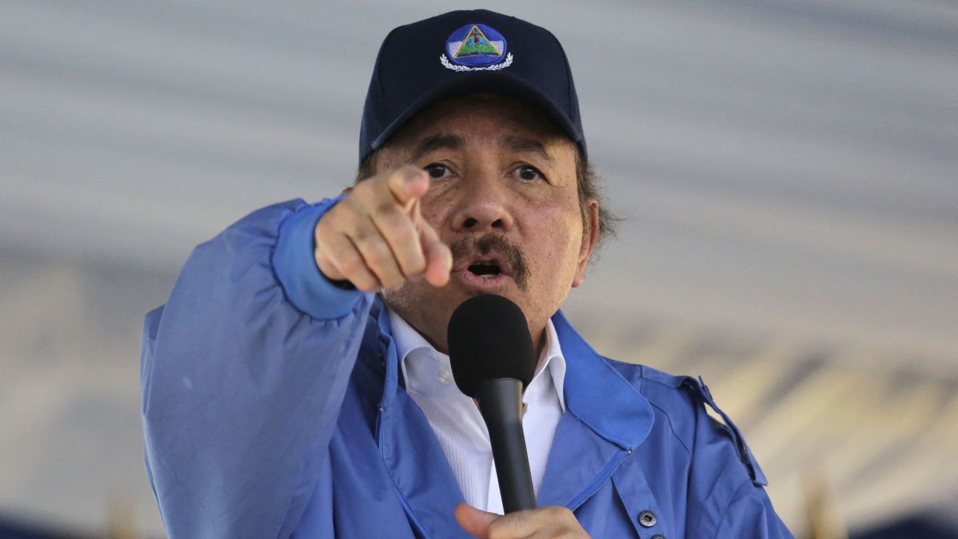 Nicaragua president Daniel Ortega holds a microphone to his face while pointing at a crowd. 