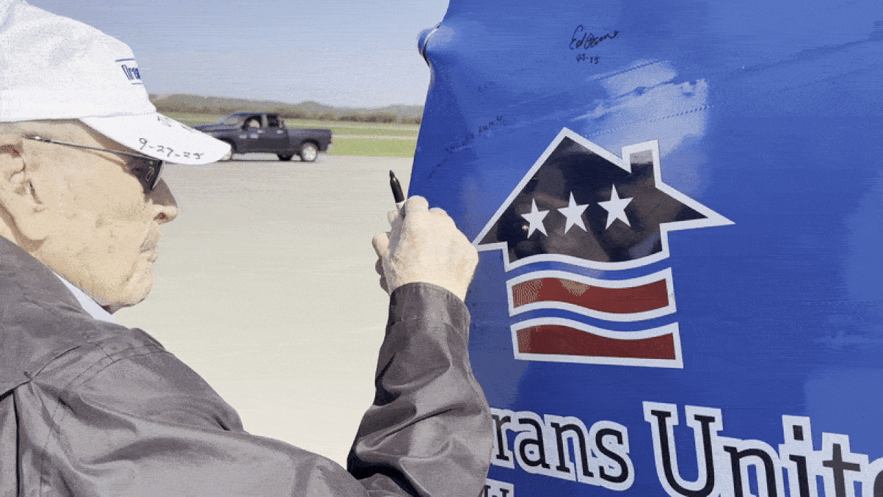 An elderly man wearing a white cap and glasses signs his name with a black marker on a blue surface featuring a house logo with three stars and red stripes. A black pickup truck is in the background.