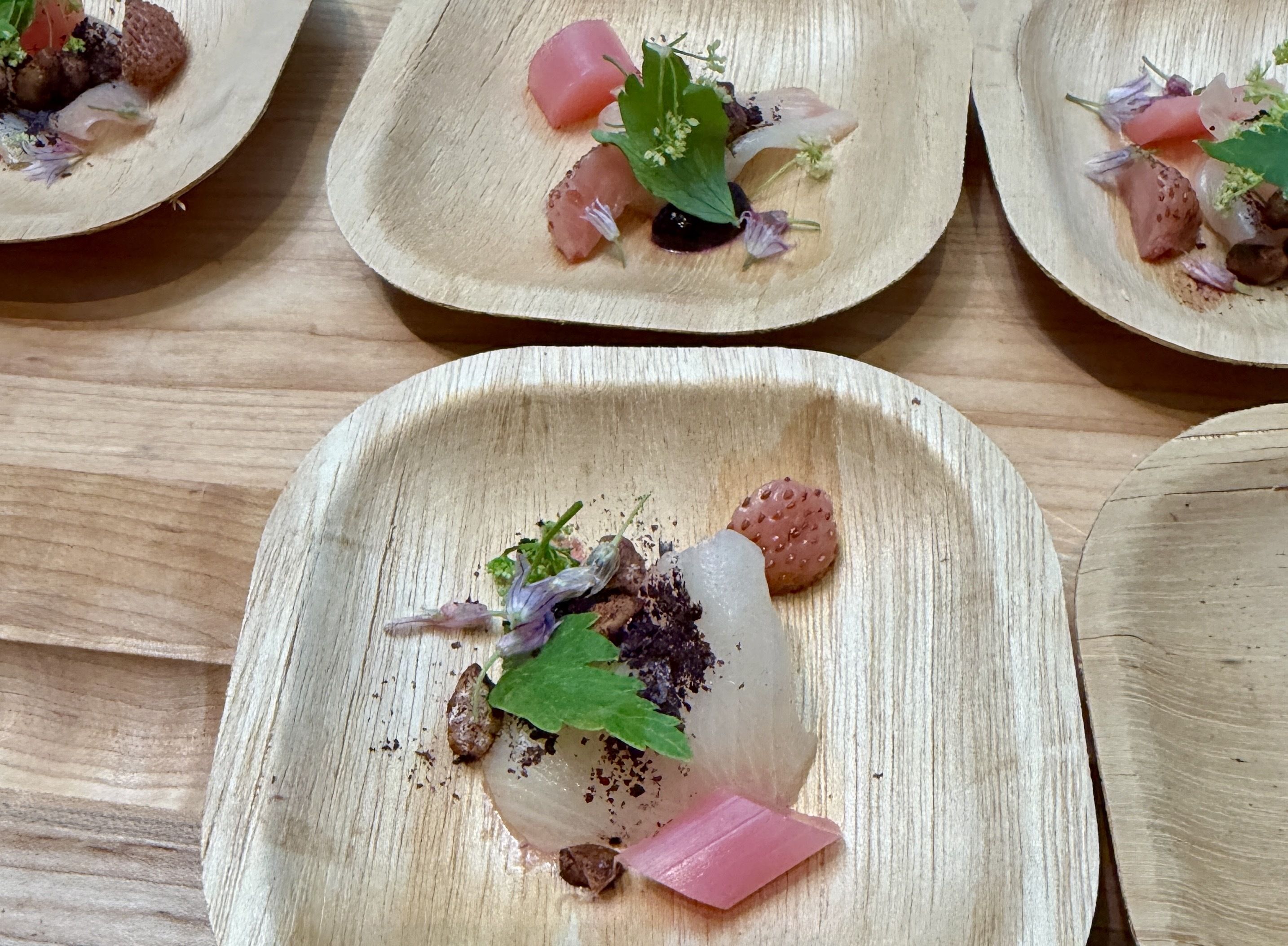 A row of light wooden serving boats on a wooden table, each topped with pink meat or fish cubes, bright green herbs, purple flower petals, and a small dab of dark sauce.