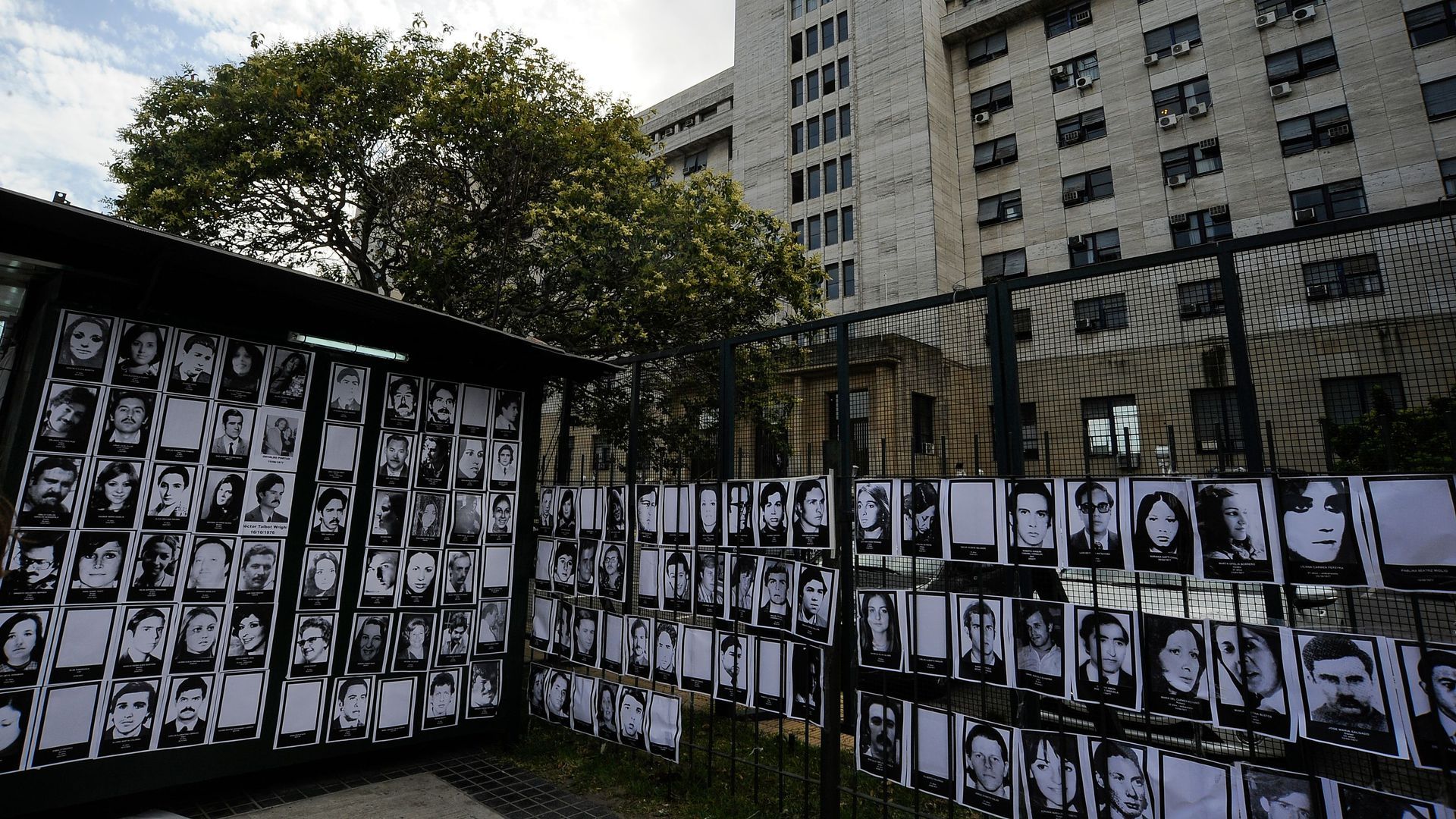 A wire fence with a grid black and white photos in front of a tall building