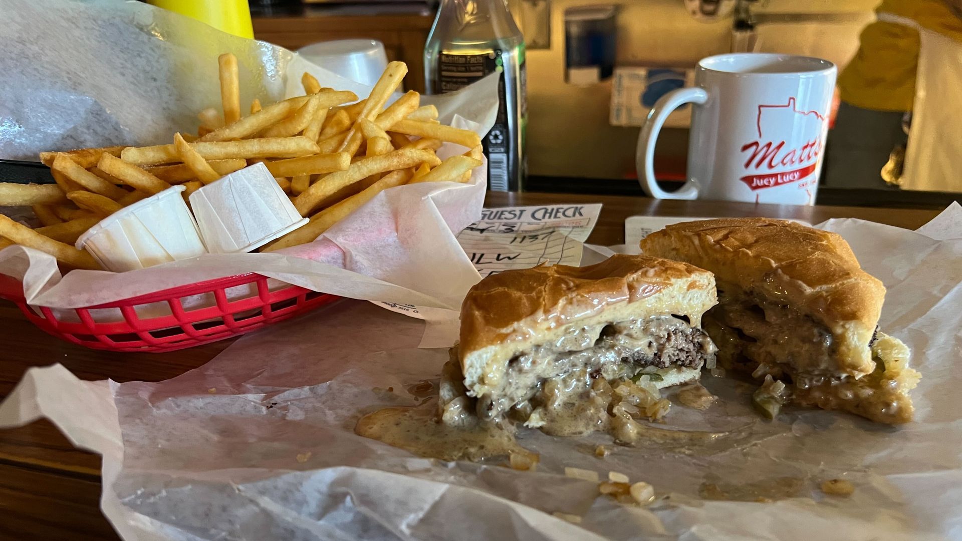 A hamburger with cheese stuffed into the middle of the patty on a bun, sliced in half to show the cheese oozing out, next to a red basket of french fries. A mug reading "Matt's Bar" in the background.