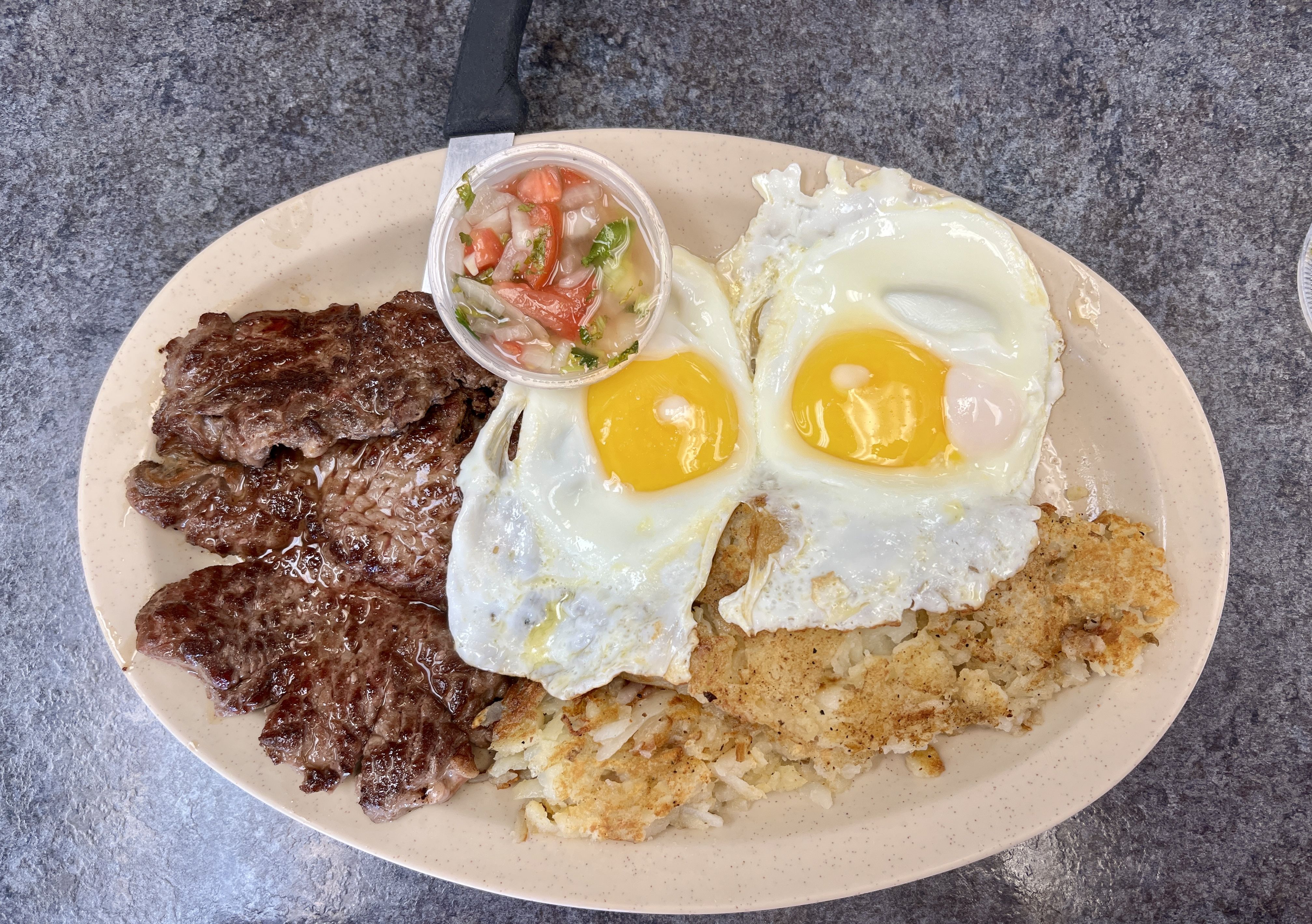 Plate with fried steak strips, two sunny-side-up eggs, crispy hash browns, and a small cup of tomato, onion, and cilantro salsa on a beige oval plate on a gray countertop.