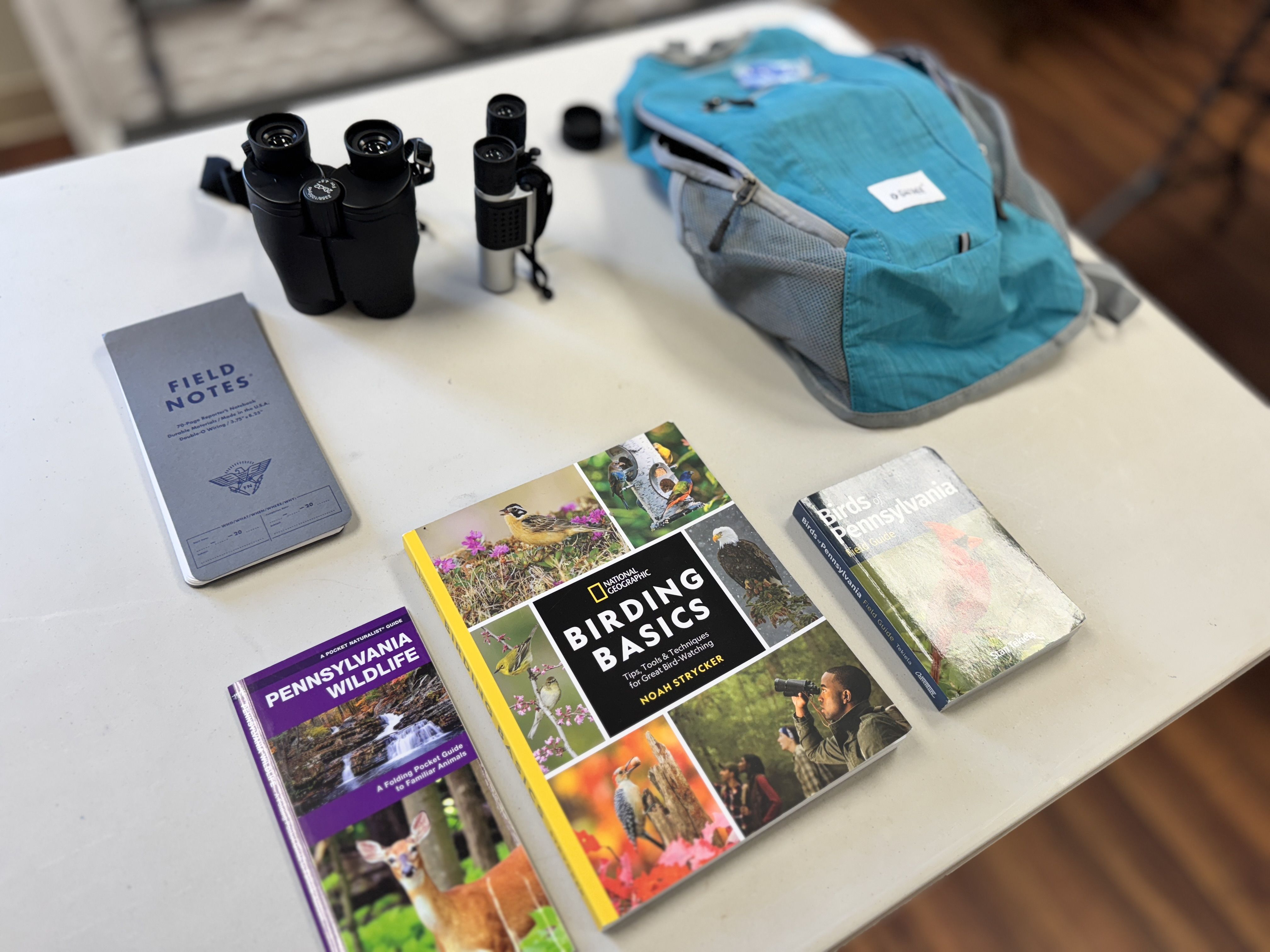 On a white table sits a teal backpack beside black binoculars and a gray field notes notebook; nearby are colorful bird and nature guides, including National Geographic titles.