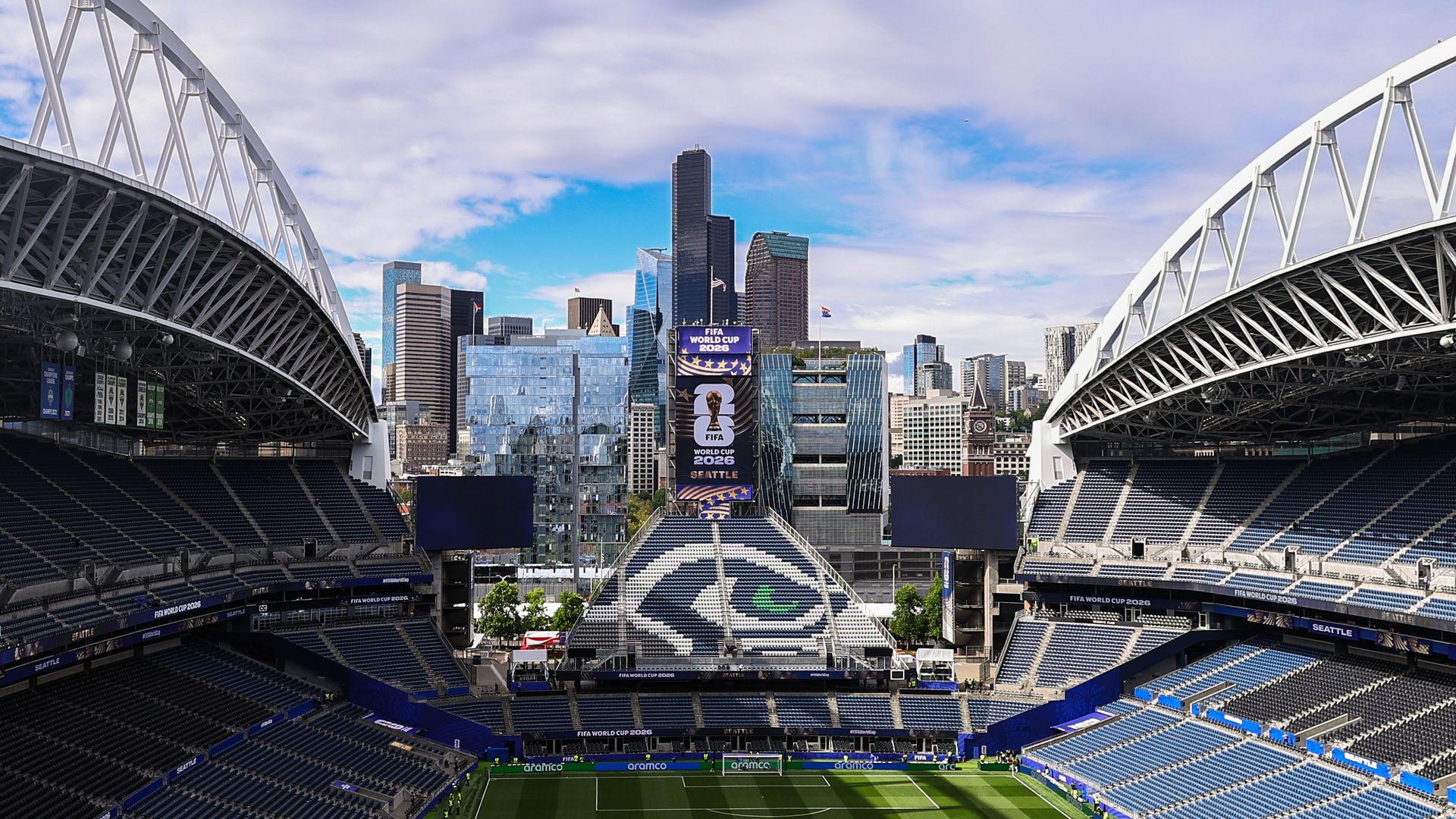 A view of an empty stadium with a city skyline behind it and a Seahawks logo on the seats in one central part of the stadium. Blue sky and some clouds overhead.