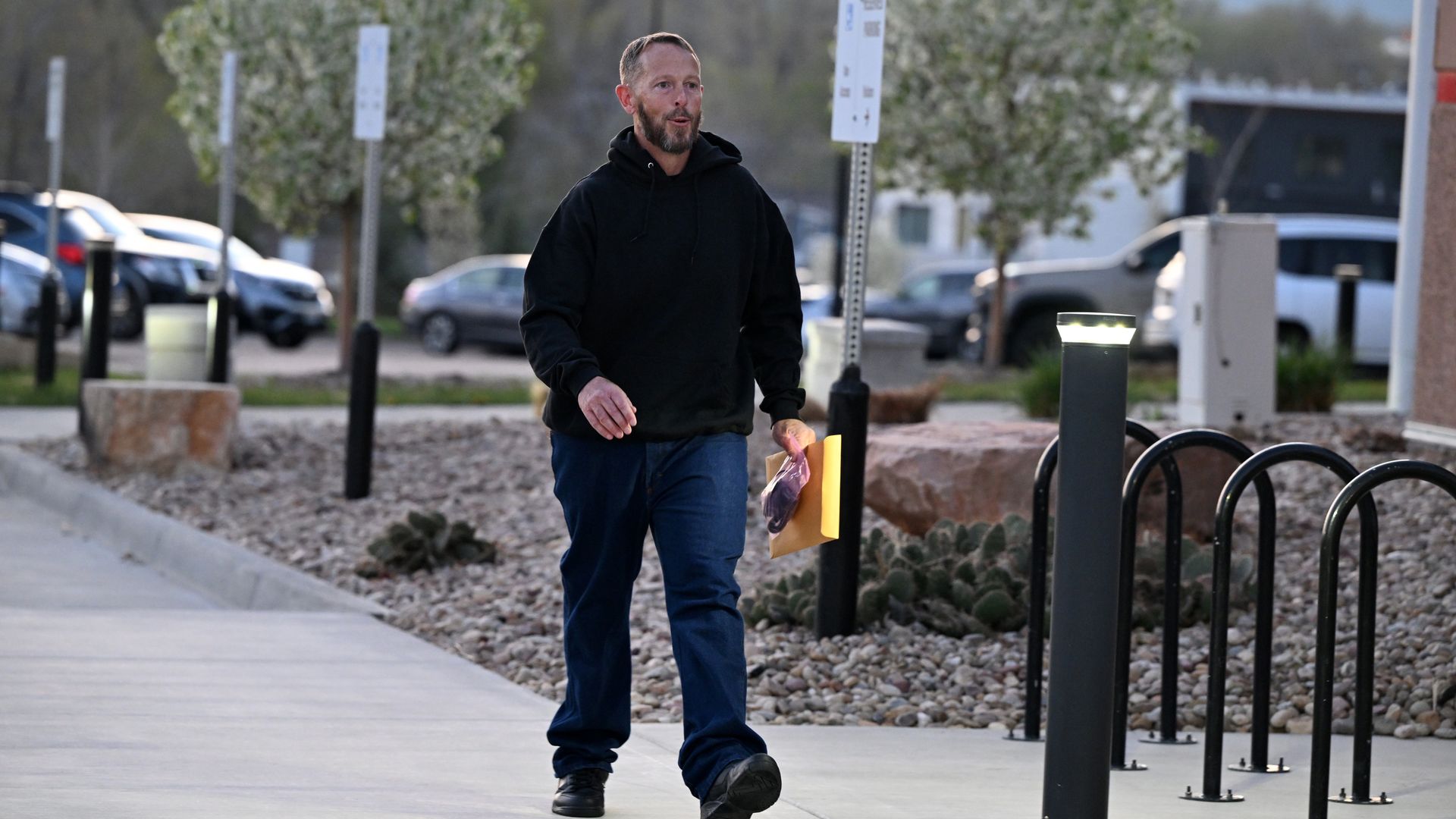 Michael Clark walks in a black sweatshirt and holding an envelope and bag.
