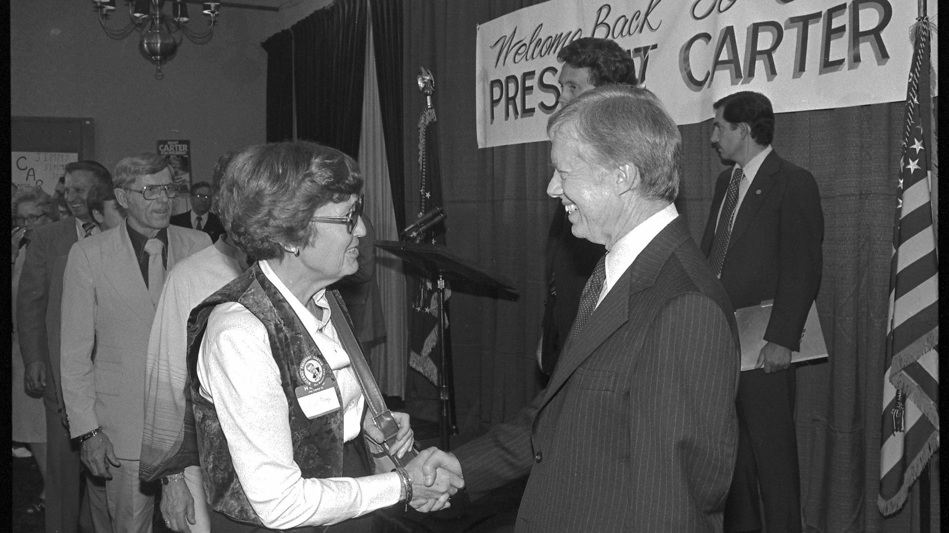 Black and white photo of Jimmy Carter shaking hands with a woman in a vest, with a sign in the background that reads "Welcome back President Carter"