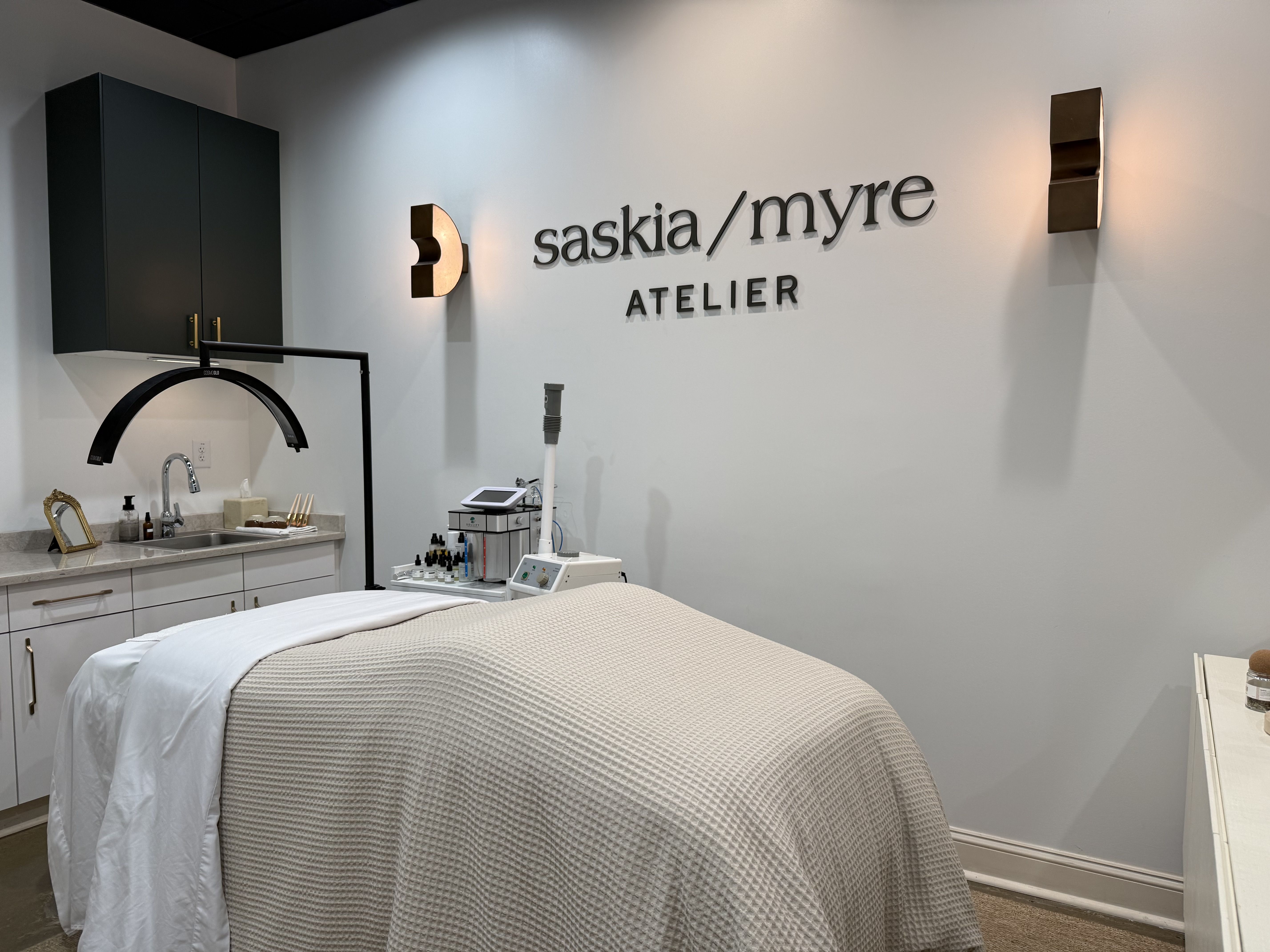 Treatment room with a beige-covered bed, sink and dark cabinets on the left, and a pale wall displaying the sign "saskia / myre" ATELIER in bold letters with warm lighting.