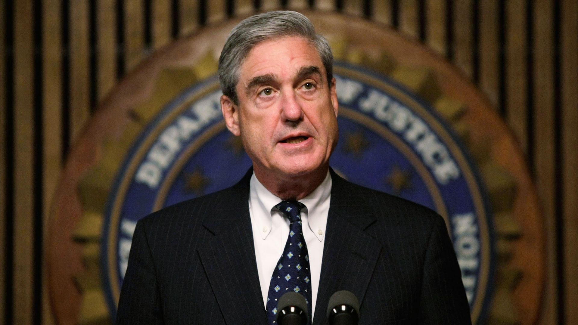 In this image, a suited Robert Mueller stands in front of a microphone podium with the logo of the Department of Justice behind him.