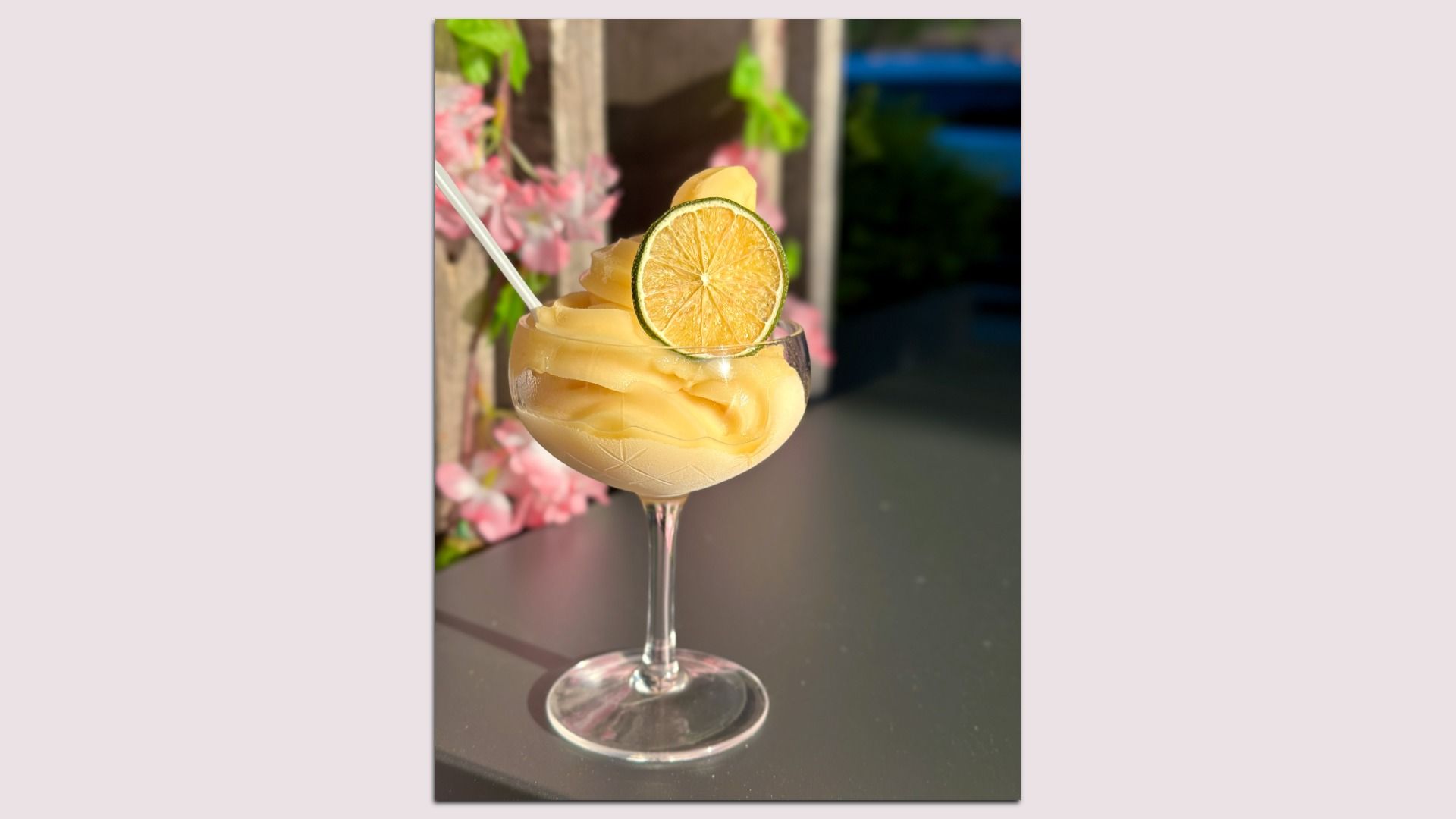 Frozen yellow cocktail in a stemmed glass with a lime wheel garnish; a white straw rests in the drink, pink flowers and greenery blurred in the sunny outdoor background.