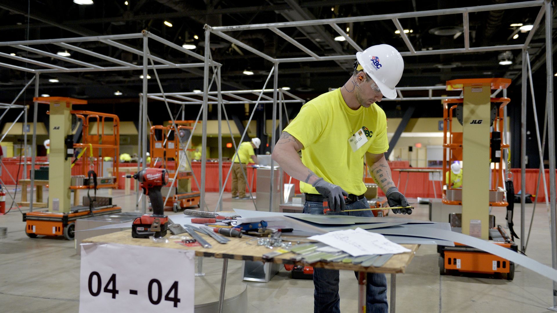 In this image, a man in a bright T-shirt and a white hard hat works over a table in a warehouse. 