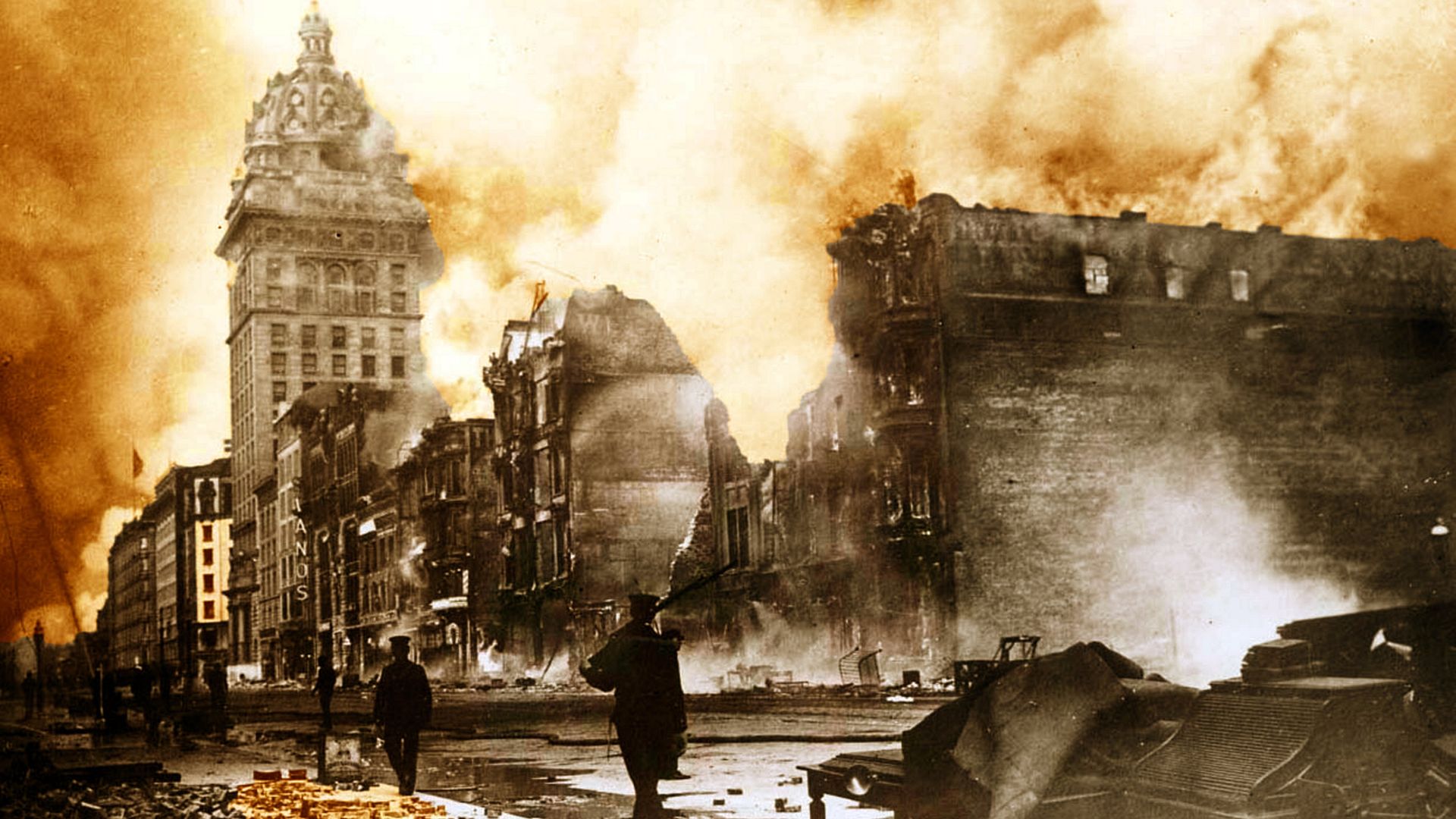 Sepia-toned photograph of a city street burning, an ornate tall building with a domed top on the left; flames and smoke rise as rubble and silhouettes of people move through debris.