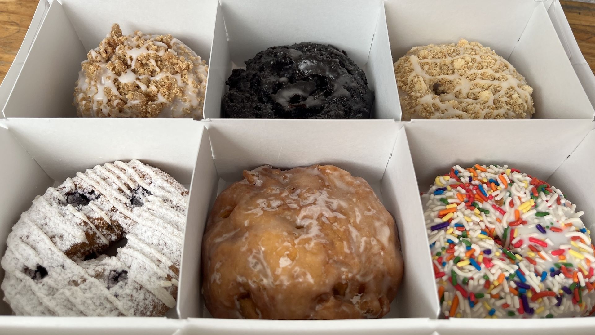 Open white bakery box with three donuts in a row: left white icing with crumb topping, center glossy dark chocolate, right crumbly glaze with white drizzle; additional donuts with powdered sugar and sprinkles visible.