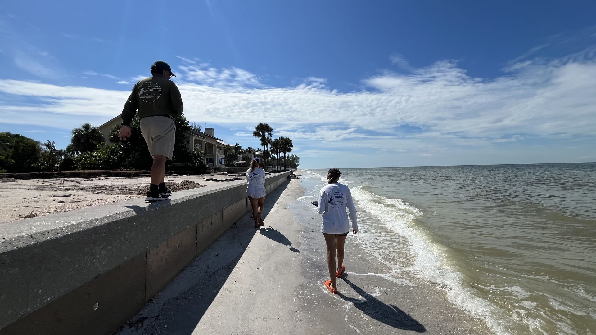 Three people walk along a narrow beach. 