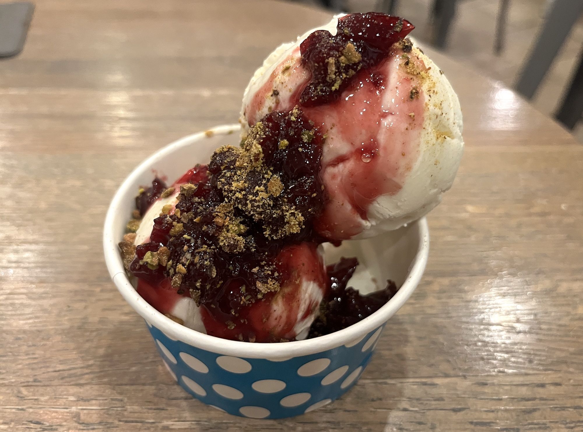 Close-up of vanilla ice cream scoops with red berry sauce and crumbly topping, served in a blue polka-dot cup on a wooden table.