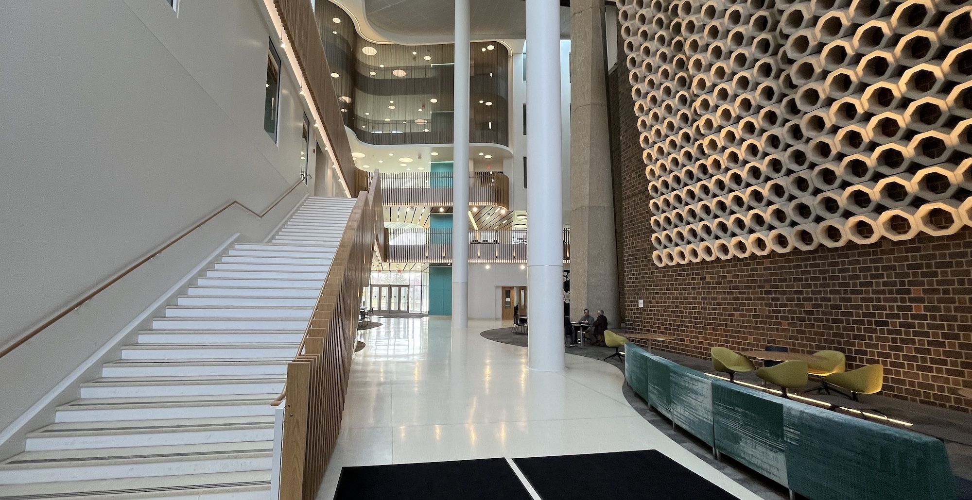Modern atrium with a long white staircase on the left, tall white columns, and glass balconies. Right wall features a honeycomb brick pattern; curved green seating with yellow chairs and a few people.