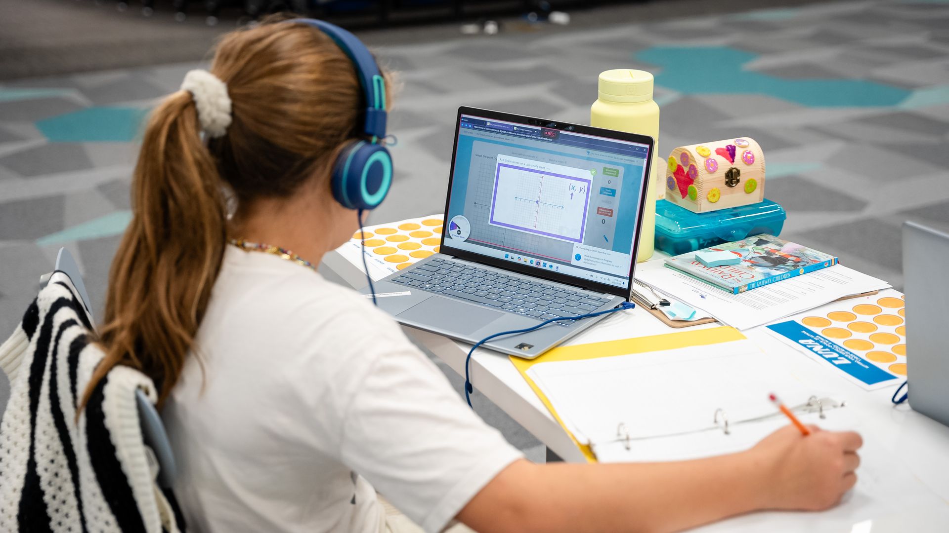 Person with long light-brown hair wearing blue over-ear headphones sits at a desk, using a laptop that shows a graph. A yellow water bottle and a colorful gift box sit nearby on a patterned floor.