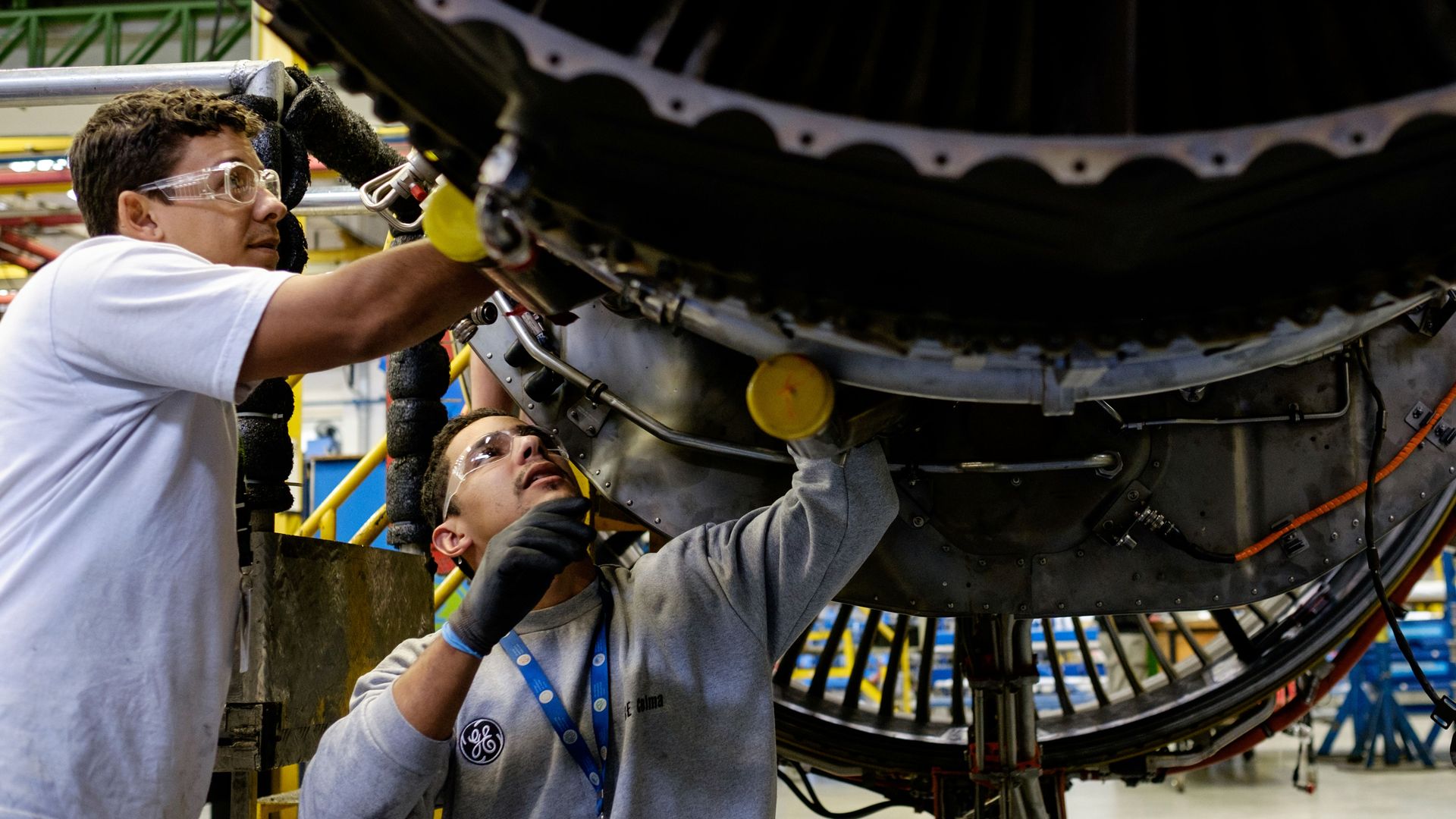 Two technicians wearing safety goggles inspect a large jet engine in a factory. A man in a white shirt on the left and two others work around the turbine with yellow scaffolding and blue lanyards.