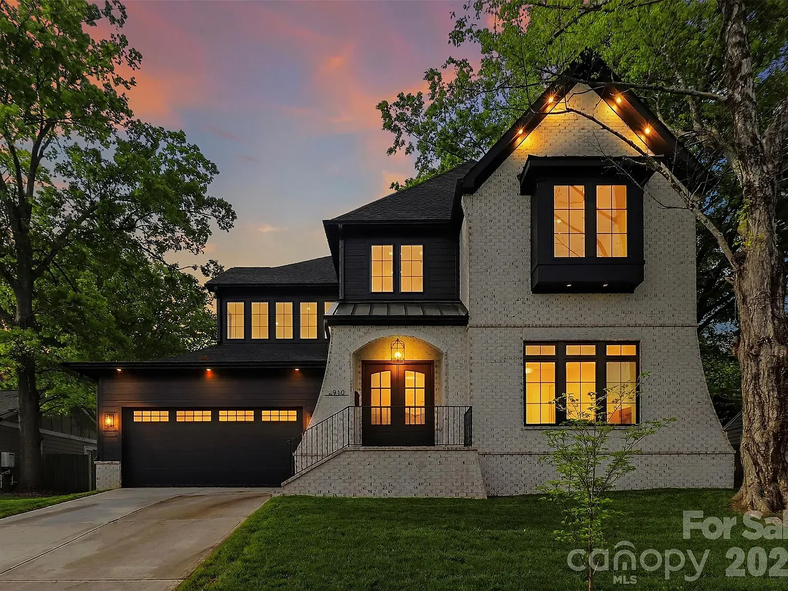 Two-story brick house with black trim and a dark garage, warm glowing windows, a small front porch, string lights on the peak, and a sunset sky with trees.