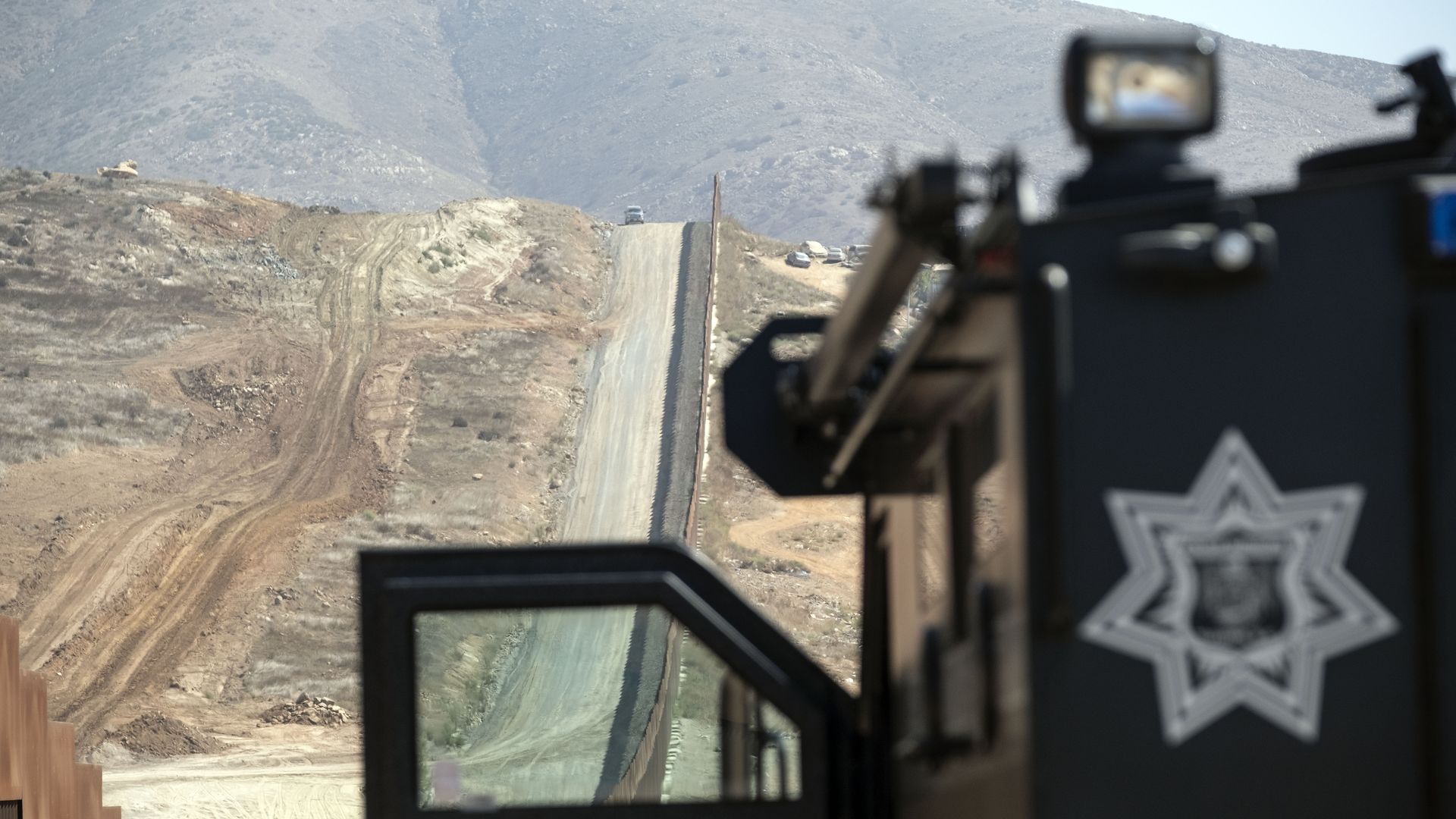 This image shows an armored truck that is facing a long border wall on a dry sandy hill