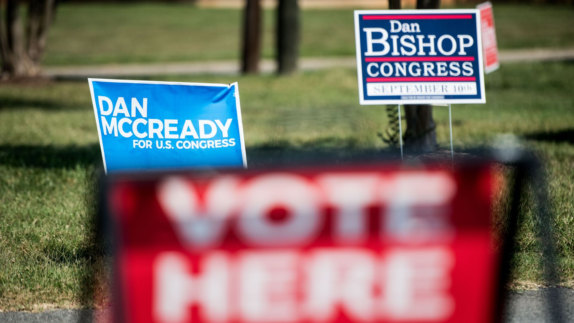 In this image, a blurred "vote here" sign rests in the foreground on a green lawn. Behind that sign, two signs stand next to each other that read "Dan McCready" and "Dan Bishop" 