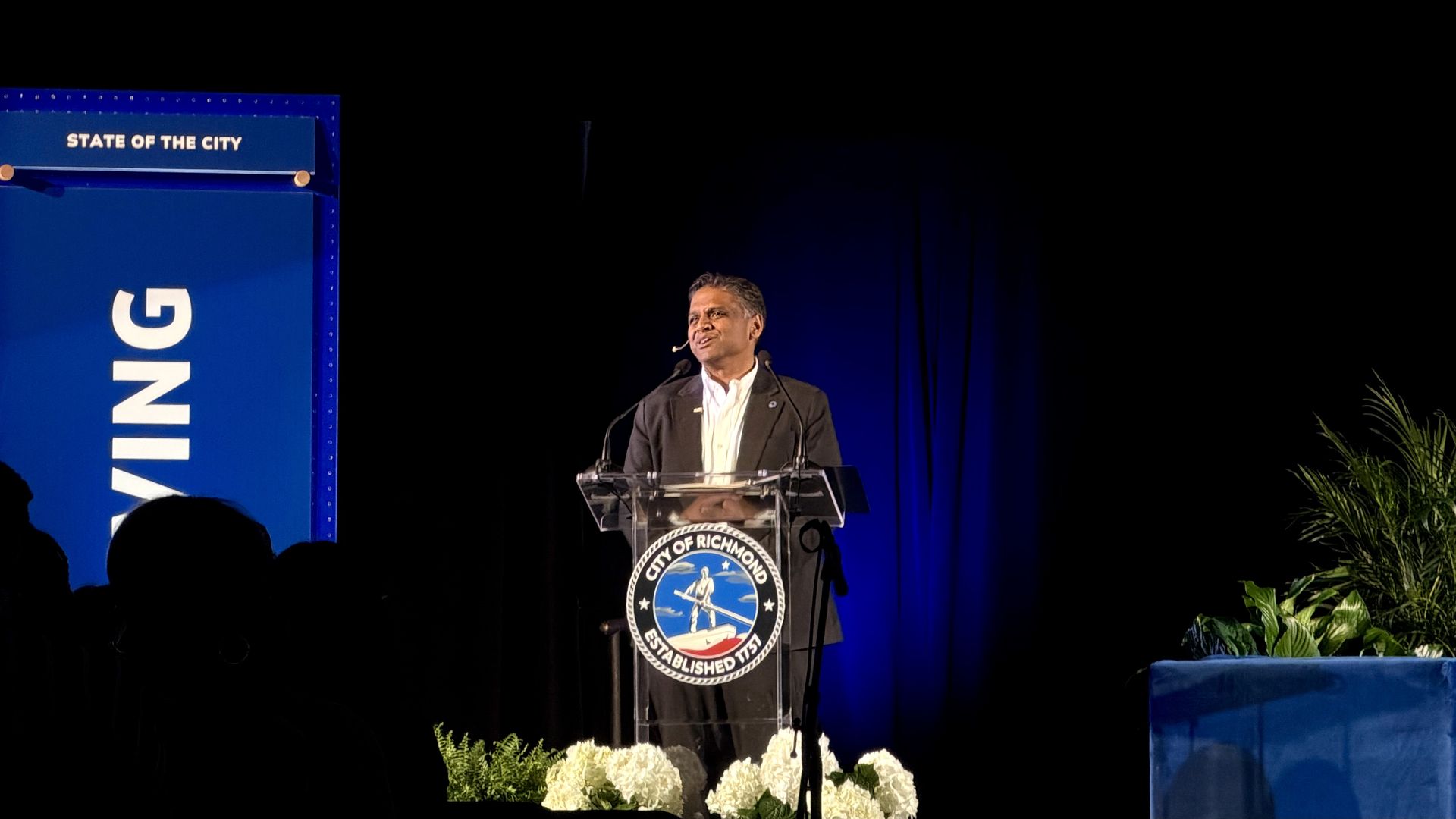Man in a dark suit speaks at a clear podium on a blue-lit stage. Left shows a blue sign reading "STATE OF THE CITY" with "RICHMOND"; audience silhouettes and white flowers in front.