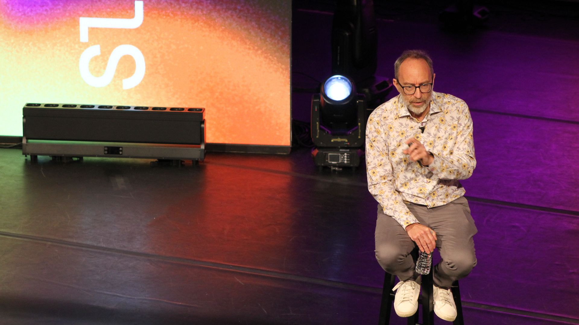 Bearded man with glasses in a floral shirt kneels on a tall black stool on a dim stage, looking down. A water bottle sits nearby, with a large orange‑to‑purple gradient screen on the left.