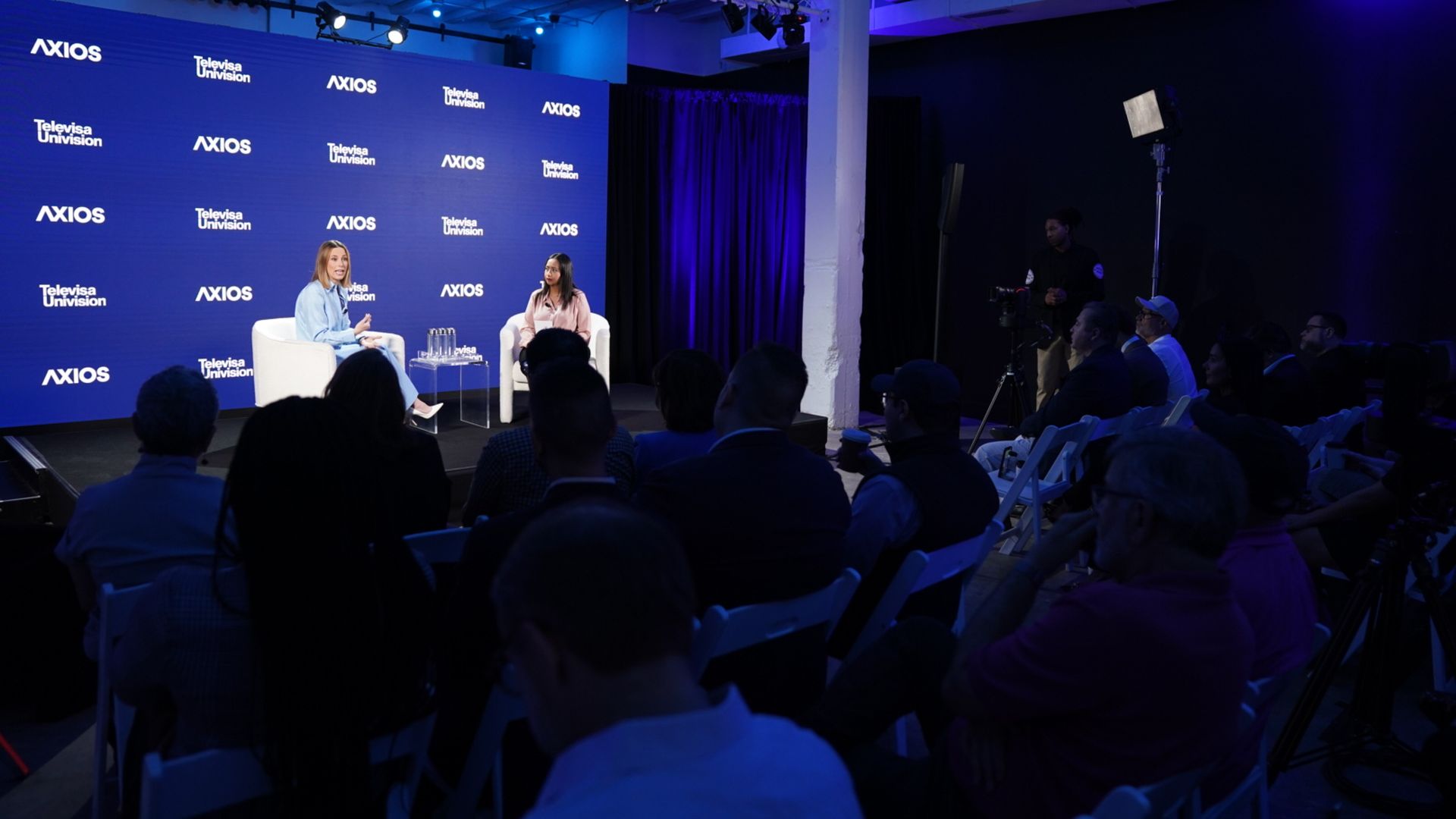 Two women sit on white chairs on a blue-lit stage for a panel, with a backdrop repeating "Axios" and "TelevisaUnivision" logos; audience watches as cameras film.
