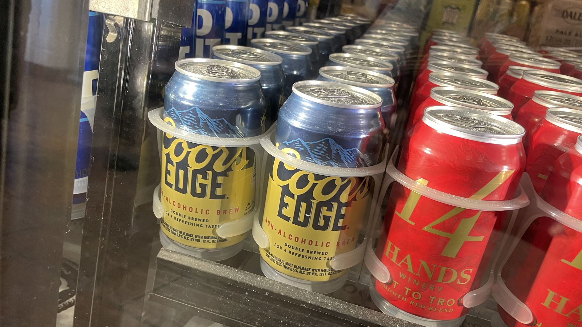 Four rows of canned beer bottles, with one reading COORS EDGE, in a blue and yellow can, and a bright red can with the words 14 HANDS on it. All the bottles are behind a glass refrigerator door. 
