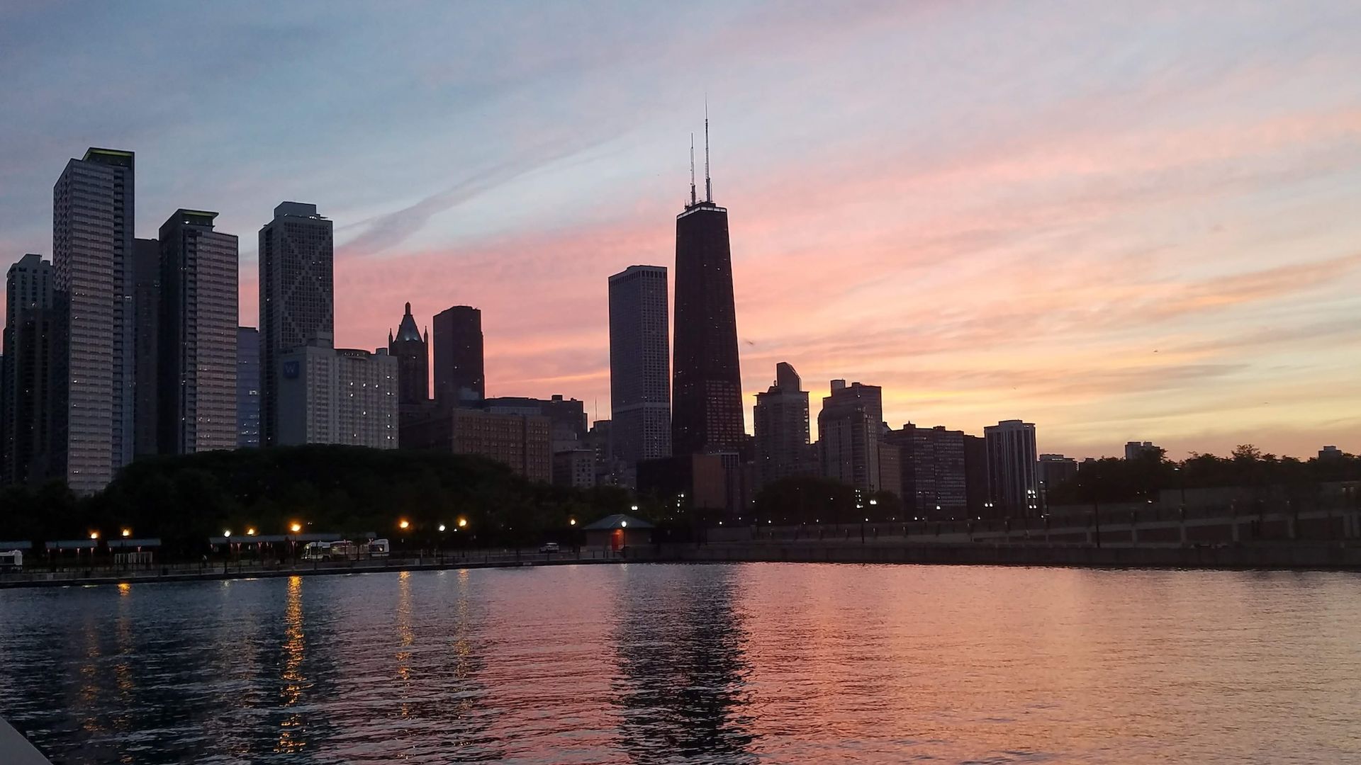 Dusk city skyline along a calm river: tall buildings on the left, a dark central tower with antennas, and a pink-blue sunset sky reflected in the water.
