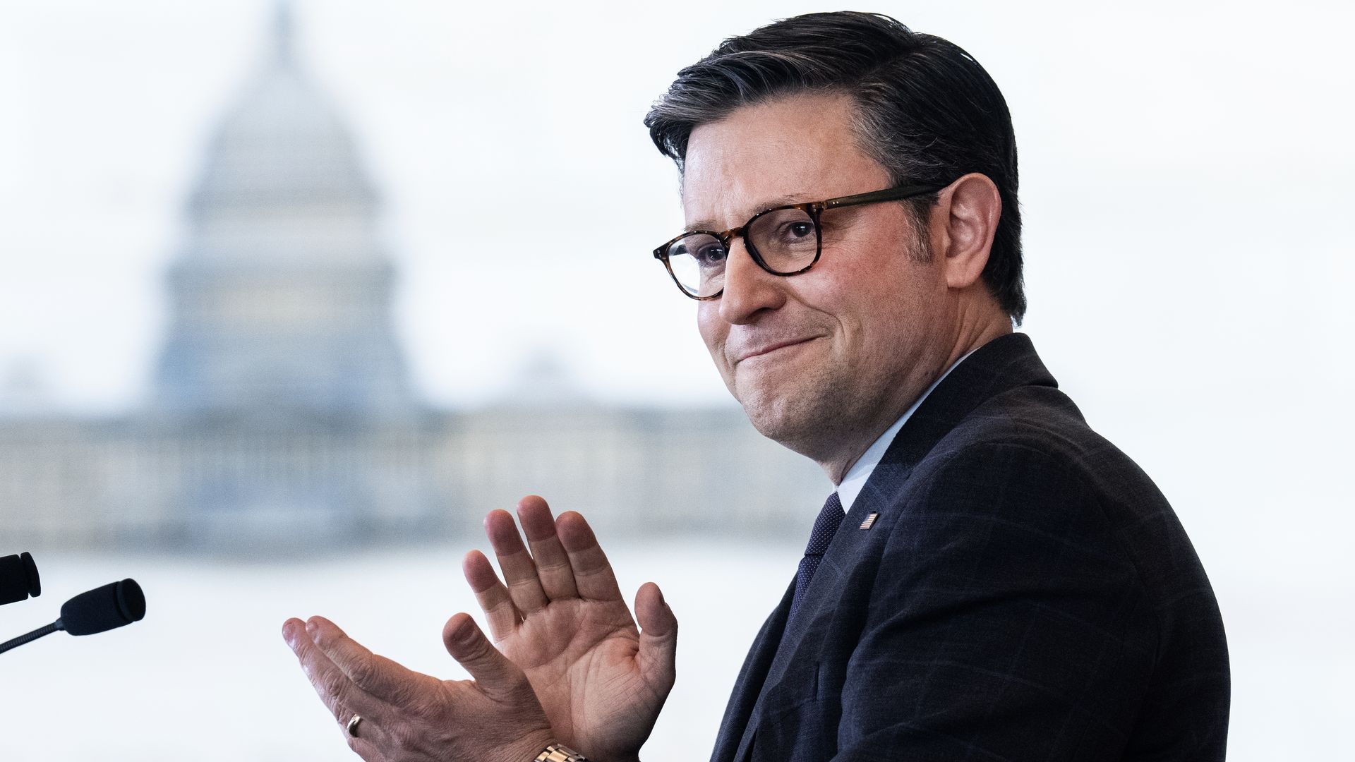 House Speaker Mike Johnson, wearing a dark gray suit and light blue shirt, clapping in front of microphones.