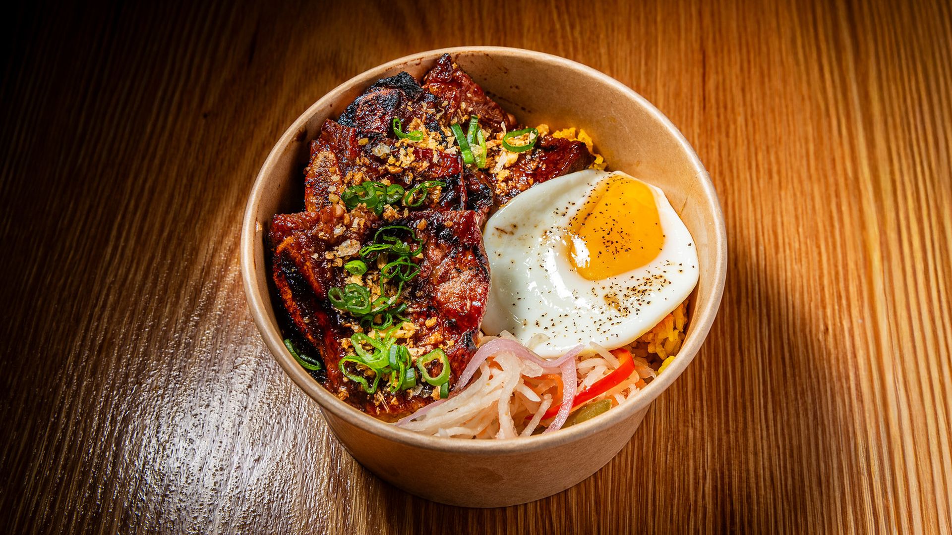 Top-down view of a brown takeout bowl of rice topped with glazed meat, a sunny-side-up egg, chopped green onions, sesame, and pickled onions, on a wooden table.