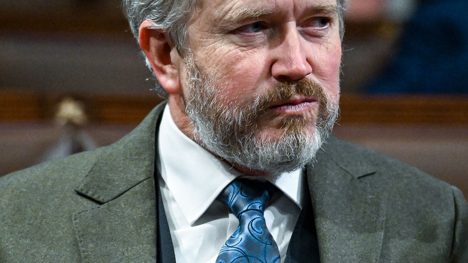 Close-up of a man with gray hair and beard in a gray suit, white shirt, and blue patterned tie, looking to the right; lapel badge reads "U.S. National Debt" with red digits.