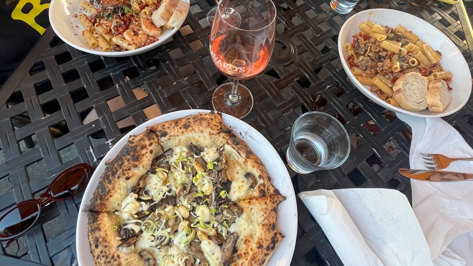 Outdoor meal on a black lattice table: a mushroom-onion pizza in a white dish, two pasta bowls (left seafood, right meat sauce with penne and bread), a glass of rosΓ©, a water glass, sunglasses, napkin and cutlery.