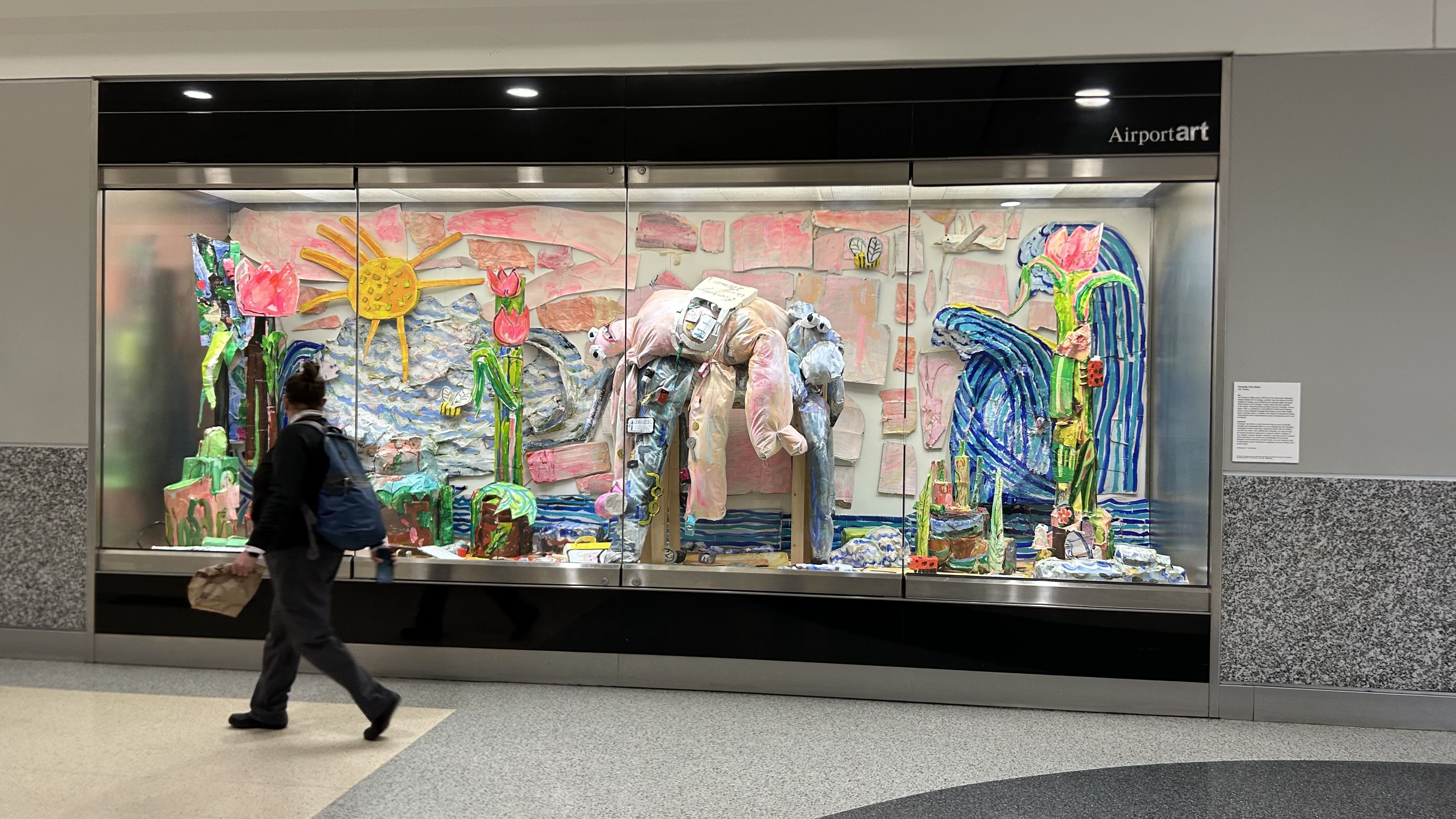 A traveler walks past a display case filled with an elaborate artwork installation of plush objects draped over furniture